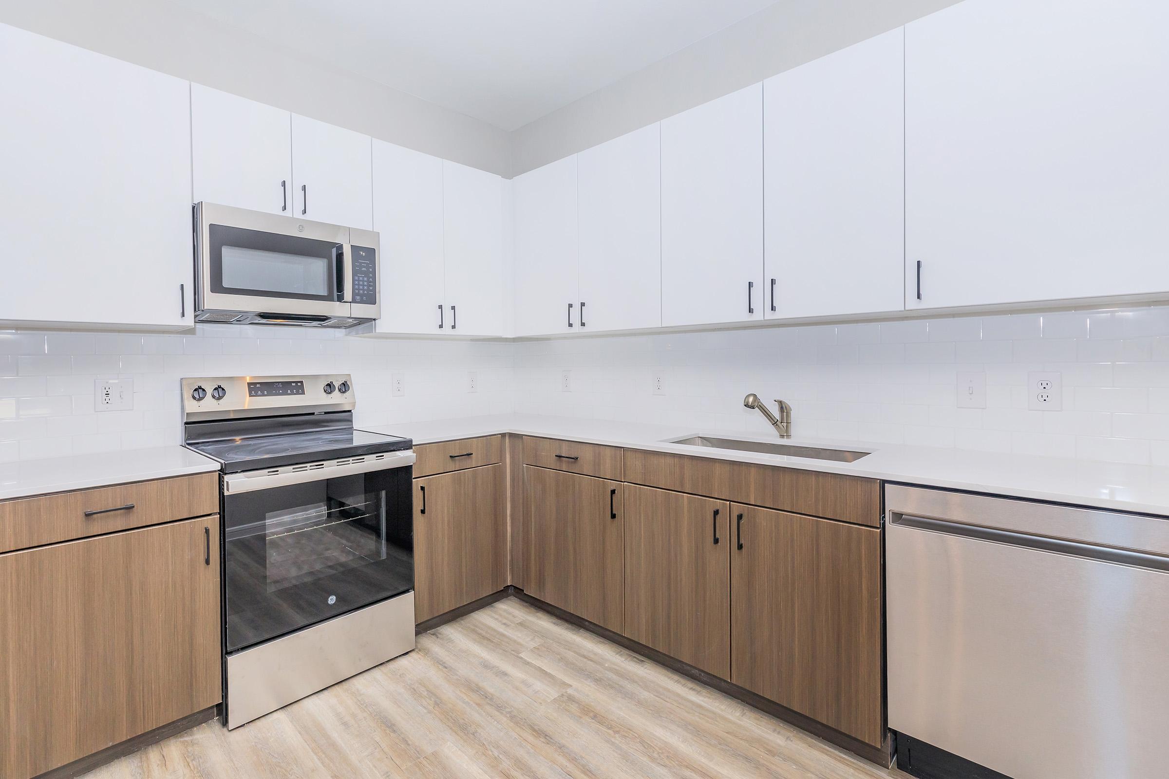 Modern kitchen featuring white cabinetry and a combination of wooden lower cabinets. It includes stainless steel appliances: a microwave, oven, and dishwasher. The countertop is a light color with a single sink, and the flooring is a light wood-like material. The overall design is clean and contemporary.