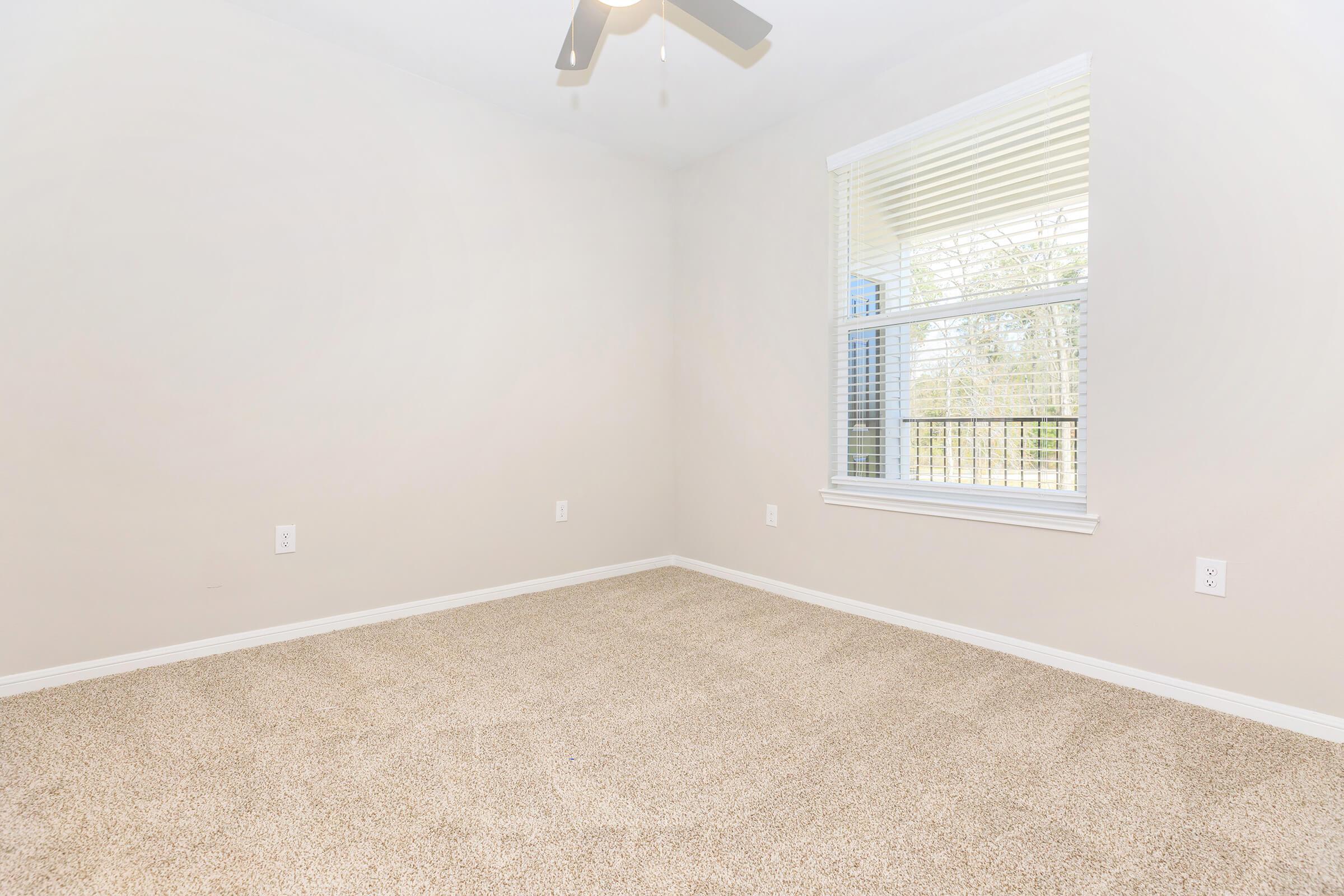 Empty room with light-colored walls and carpeted flooring. A ceiling fan is visible, and there is a window with blinds allowing natural light to enter. The room appears spacious and uncluttered, suggesting it could be used for various purposes.