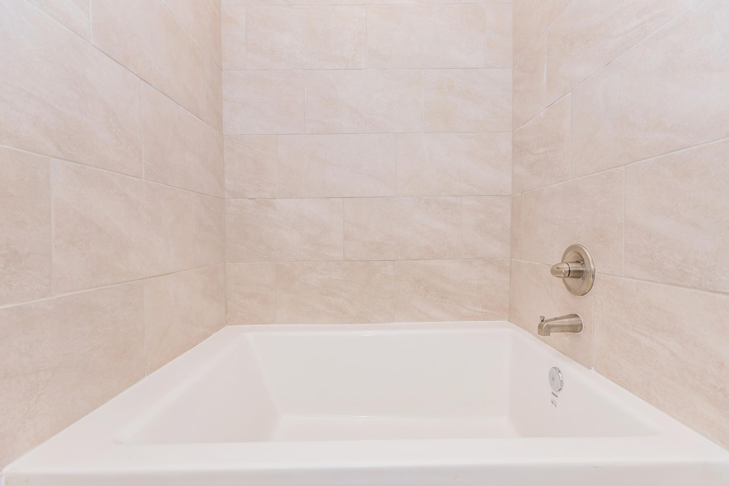 A clean, modern bathtub with a white finish, situated against light-colored tiled walls. The bathtub features a simple design and a faucet on the side, highlighting a minimalistic and spacious bathroom aesthetic.