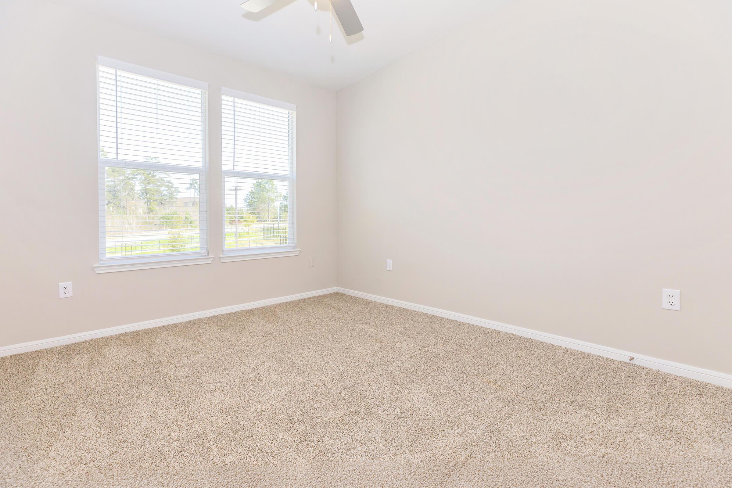 A vacant, well-lit room with light beige walls and carpet. It features two large windows with white blinds, allowing natural light to enter. The ceiling has a ceiling fan, and there are no furnishings or decorations in the space, giving it an open and spacious feel.
