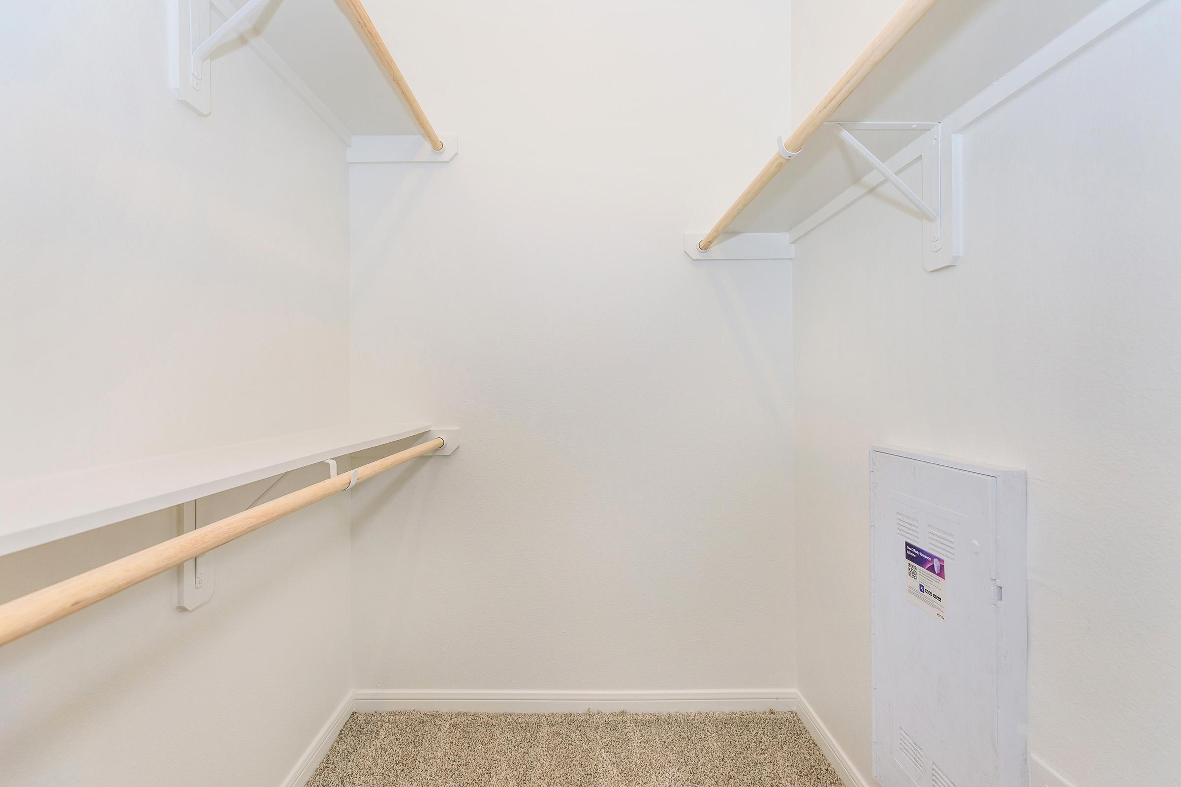 Empty closet with white walls, two wooden garment rods, and a shelf on one side. The floor is carpeted, and there is an electrical panel on the wall. The space is well-lit and organized, offering ample storage potential.