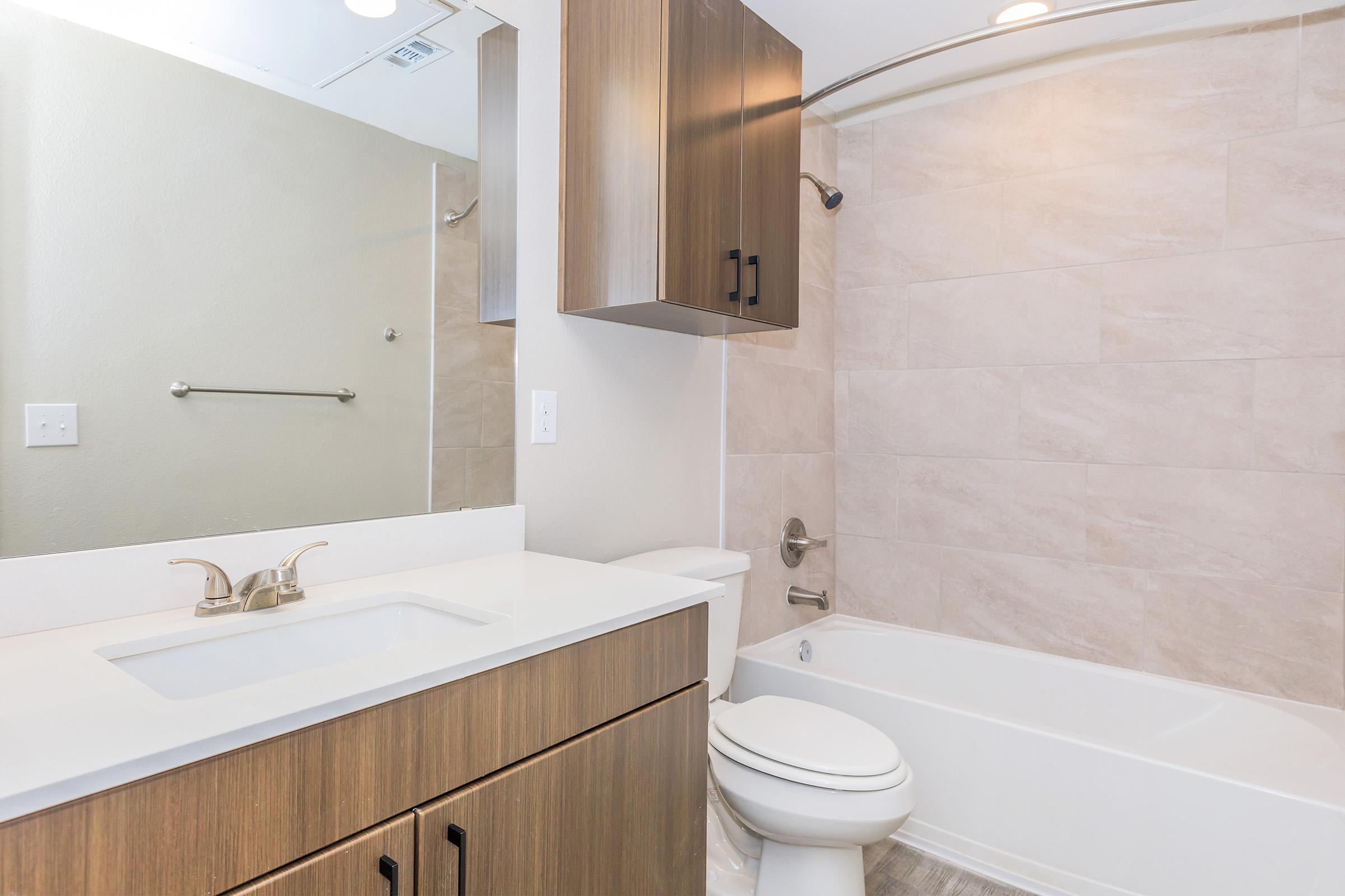 A modern bathroom featuring a white sink with a large mirror above, wooden cabinetry beneath, a bathtub with a shower curtain, and light-colored tiled walls. The space is well-lit with a neutral color palette, creating a clean and inviting atmosphere.