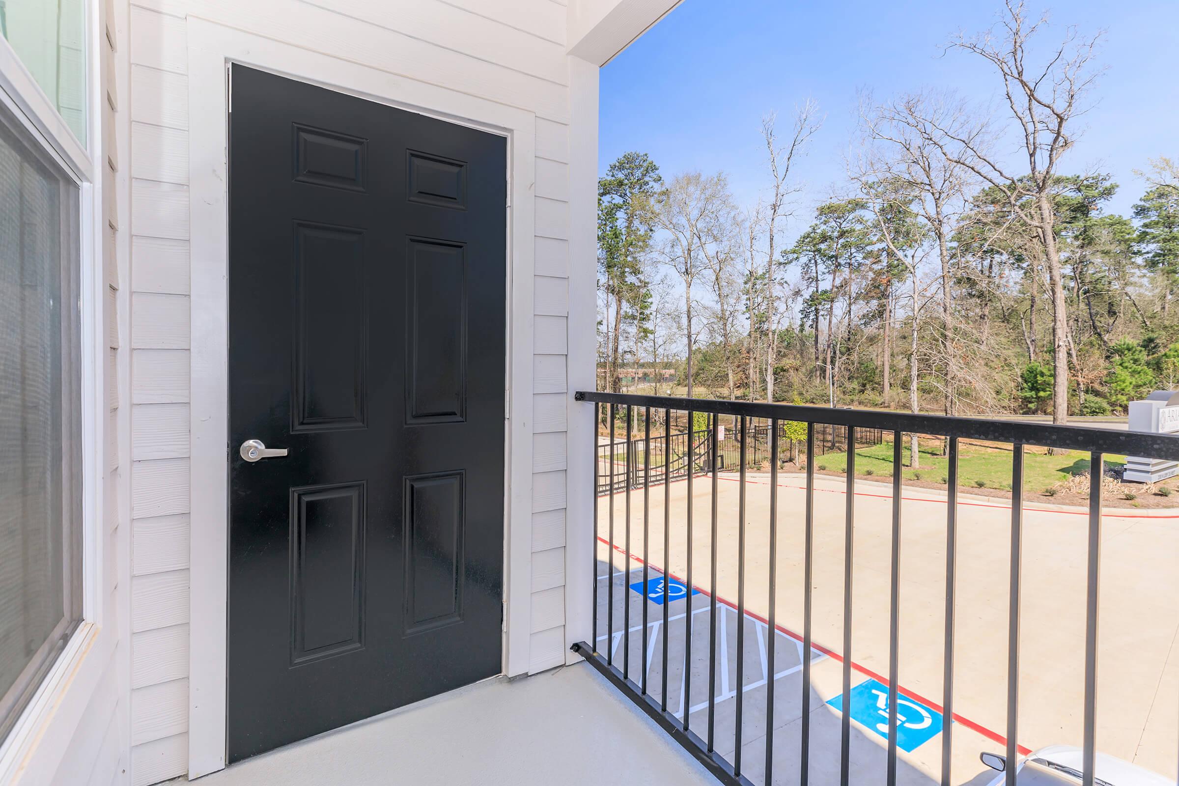 A modern balcony view featuring a black door, with a railing in the foreground. The background shows a tree-lined area outside, with a glimpse of a parking lot that has designated spaces. The scene suggests a peaceful environment with natural surroundings.