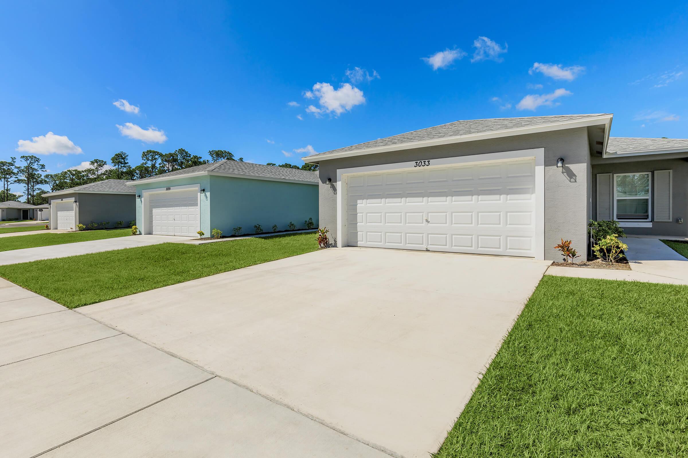 Two residential houses with light-colored exteriors and white garage doors are shown. The driveways are paved, and the well-maintained lawns are green and lush. The sky is clear with a few clouds, indicating pleasant weather. The scene conveys a clean, suburban neighborhood aesthetic.
