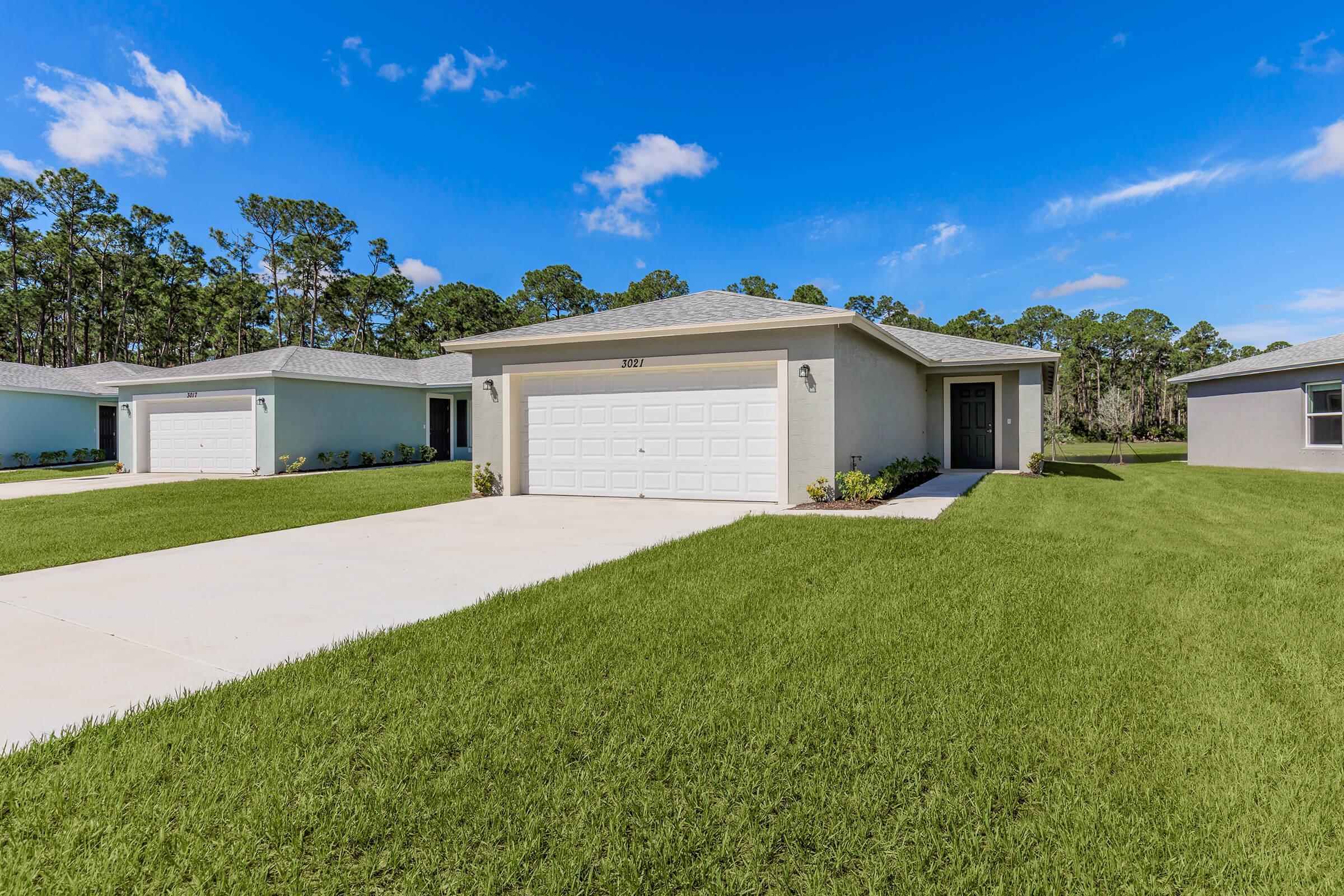 A modern single-family home with a light-colored exterior and a two-car garage. The house is surrounded by a well-maintained lawn and features small shrubs along the walkway. Clear blue skies and green trees are visible in the background, creating a peaceful suburban atmosphere.
