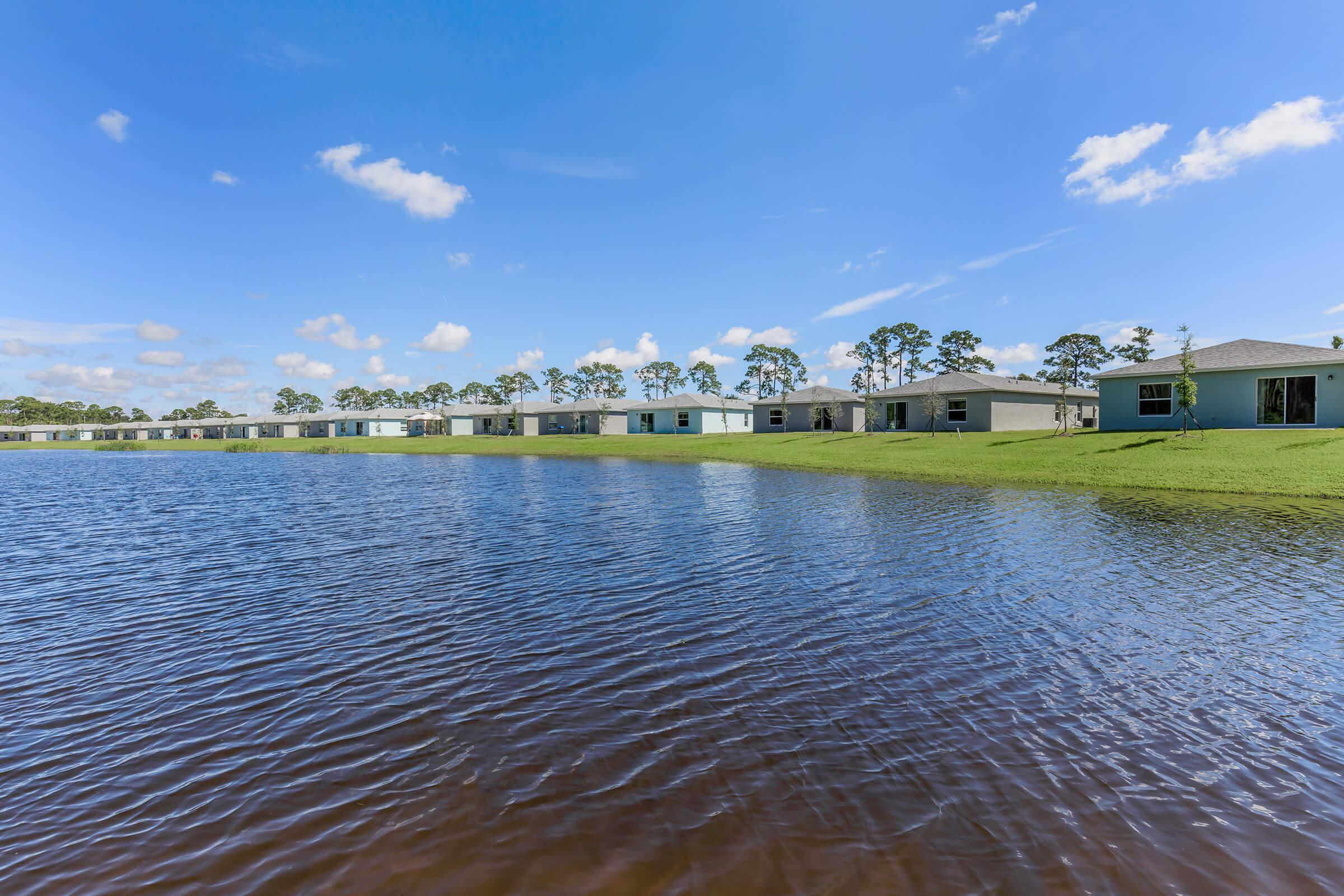 View of a calm lake surrounded by residential homes. The houses line the shore, reflecting the blue sky and fluffy clouds above. Lush green grass separates the buildings from the water, creating a serene and peaceful setting in a suburban environment.