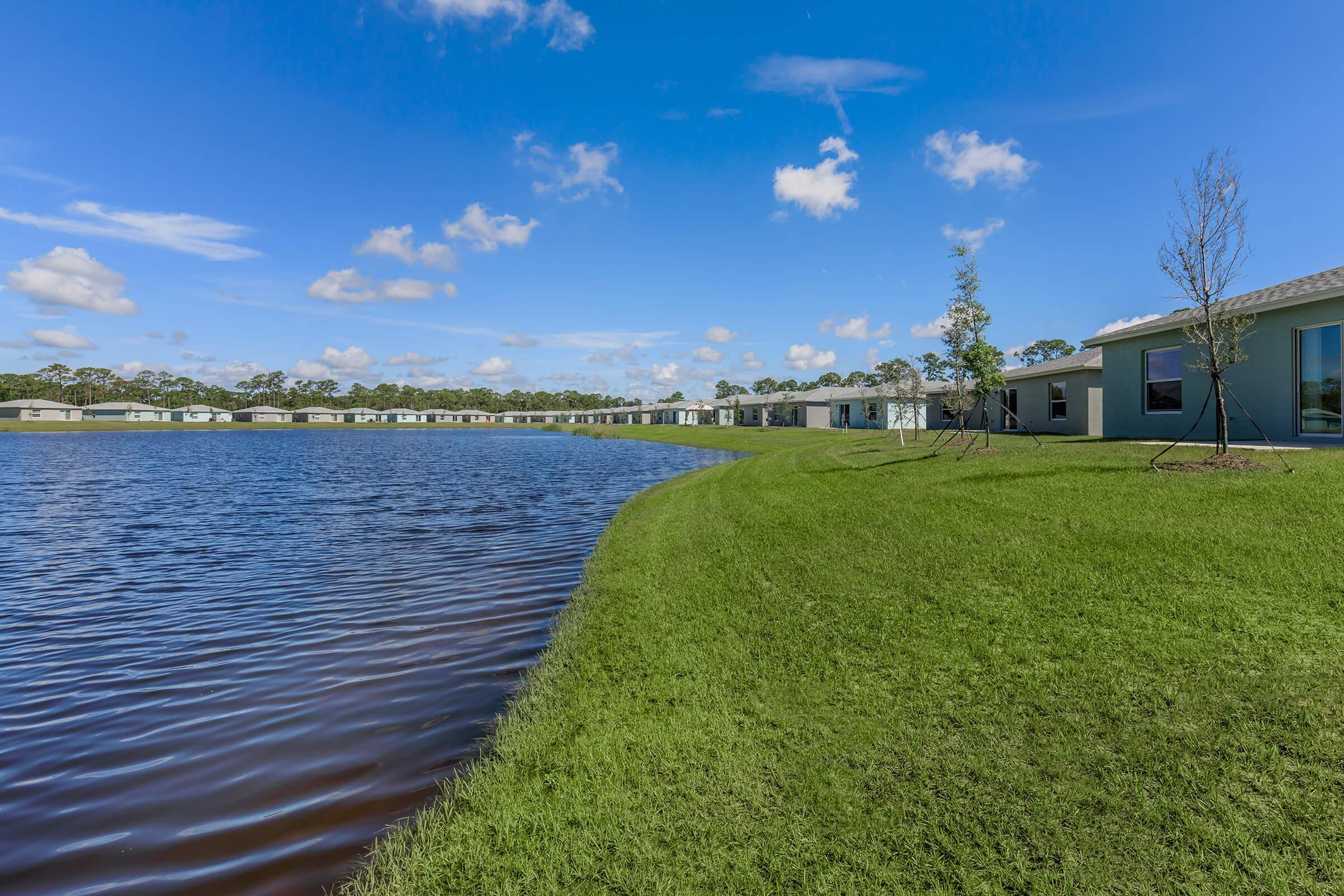 A serene view of a calm lake surrounded by lush green grass and modern residential buildings in the background. The sky is clear with a few fluffy clouds, reflecting a peaceful suburban setting.
