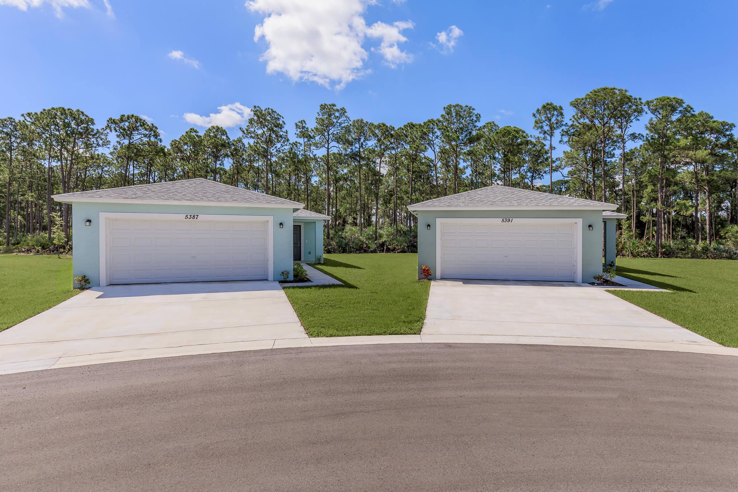 Two identical single-story houses with light blue exteriors and white garage doors, set in a serene landscape. Each house has a small porch and is flanked by well-maintained lawns. The backdrop features lush green trees under a clear blue sky, with a curved driveway leading up to the houses.