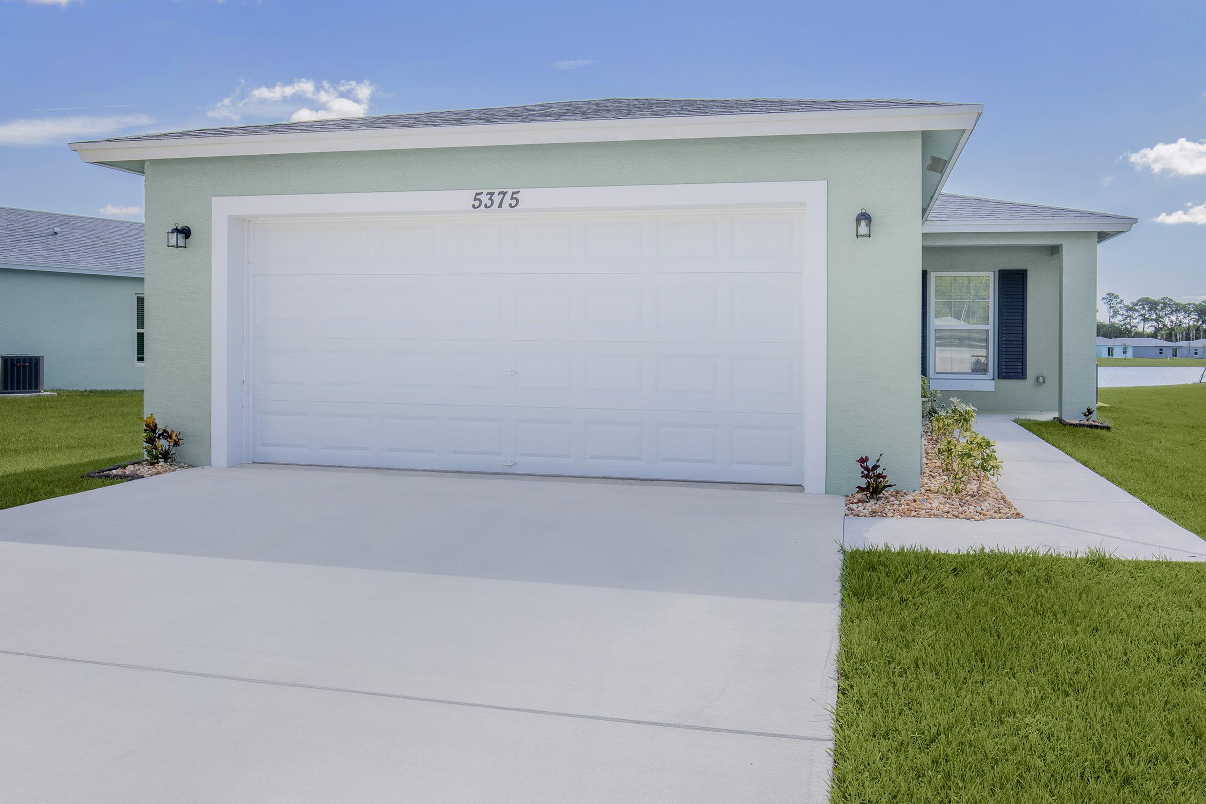 A modern light green house with a white garage door, featuring a neatly maintained driveway and small landscaping elements. The house is surrounded by lush green grass, and the sky is clear with a few clouds visible in the background.