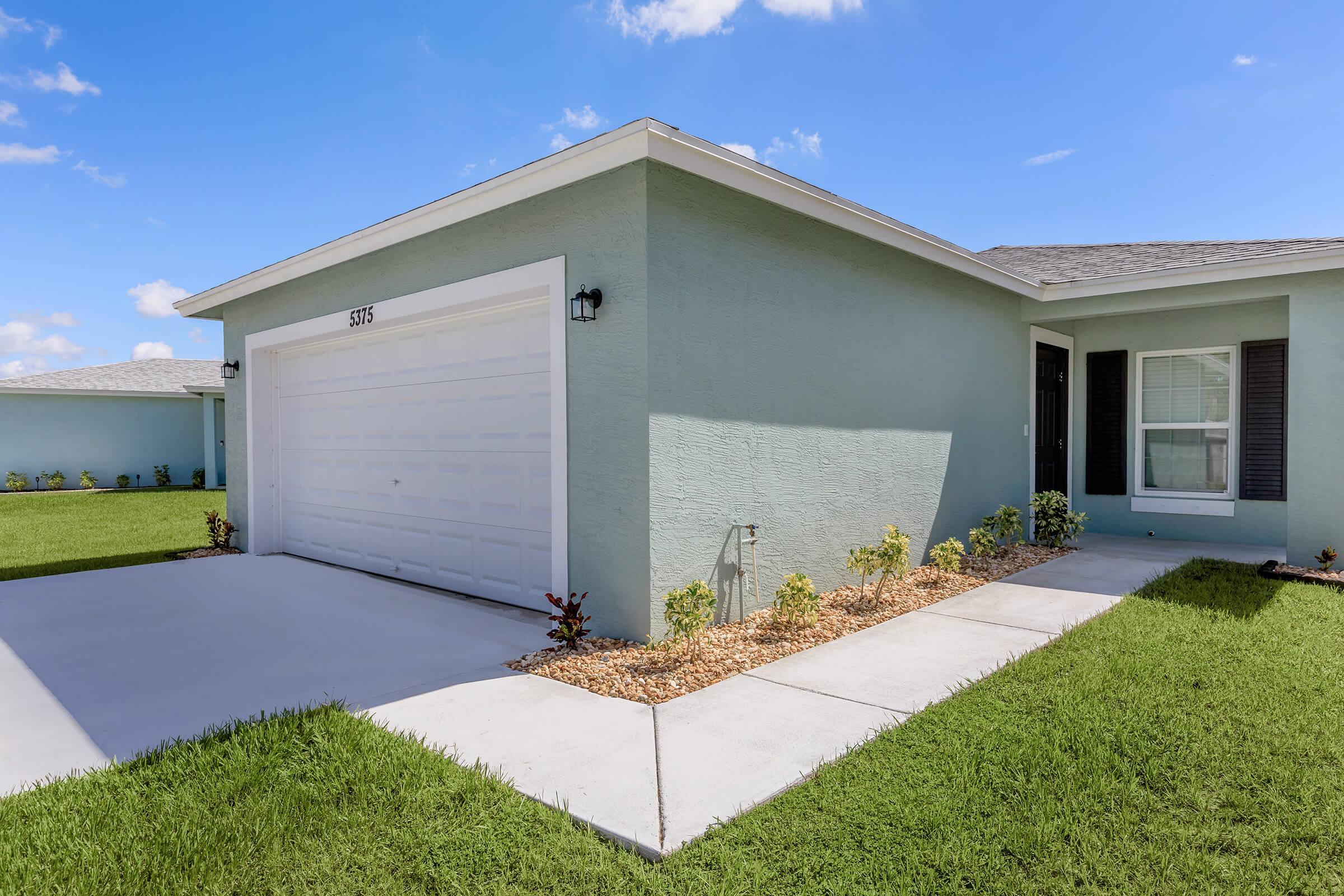A light blue house exterior featuring a two-car garage, well-maintained lawn, and flower beds along the pathway. The sky is clear with a few clouds, and the house has a modern architectural style with a welcoming entrance.