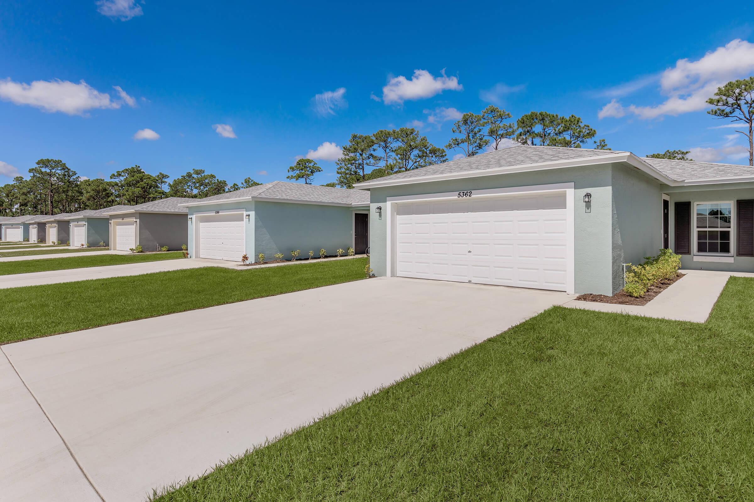 A row of modern, single-story homes with light-colored exteriors and white garage doors. The driveways are paved, and the front yards feature neatly trimmed grass. Bright blue sky with a few clouds visible in the background, giving a sunny and inviting ambiance.