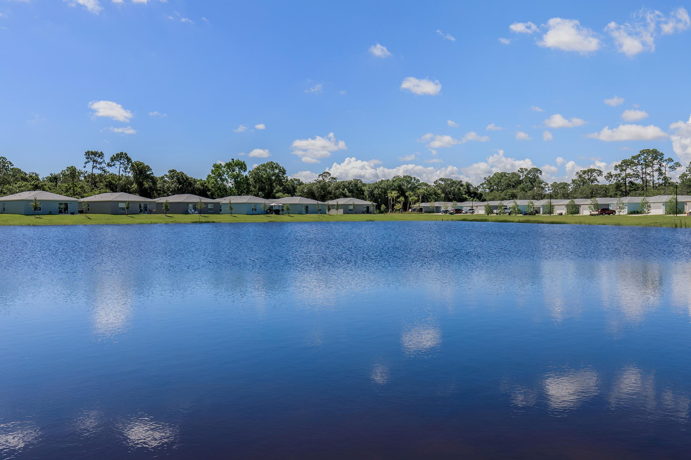 A serene lake is reflected in the clear blue water, surrounded by greenery and homes. The sky above is bright with fluffy clouds, creating a tranquil landscape ideal for relaxation and nature appreciation.