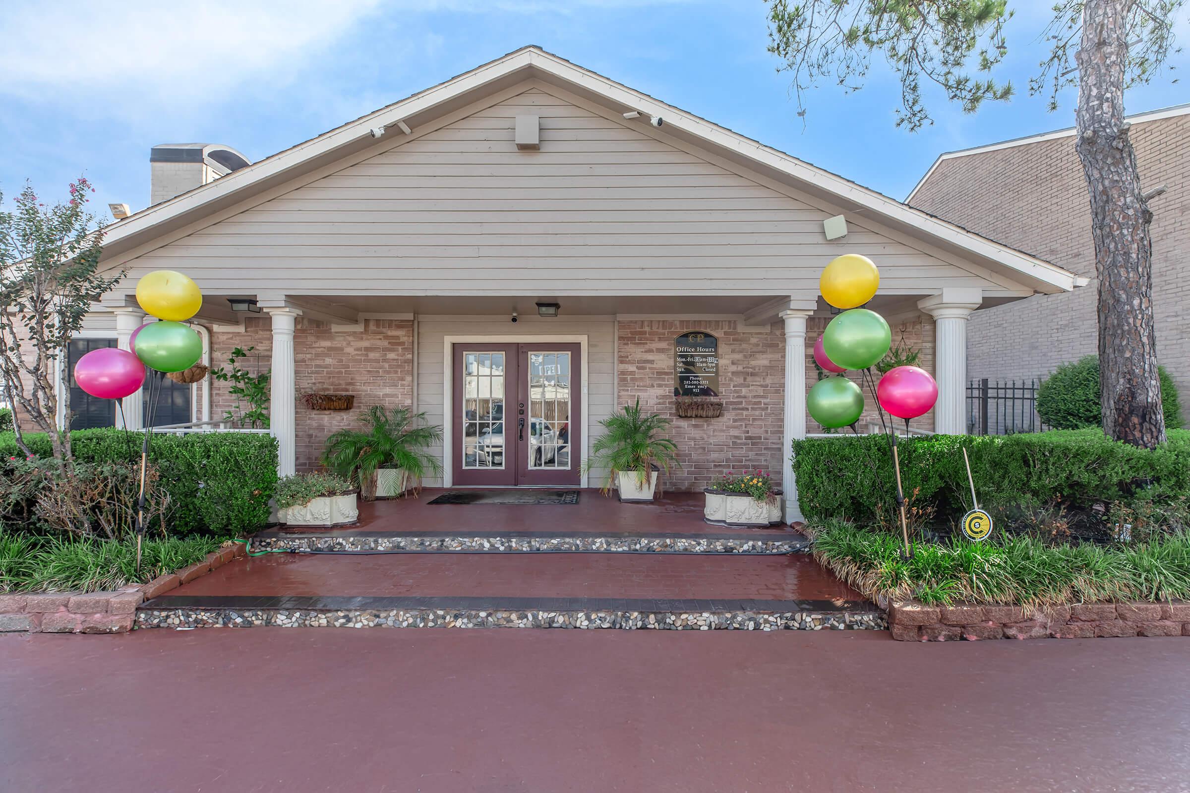 A charming entrance to a building featuring a light-colored structure with a gabled roof. Flanking the double doors are colorful balloons in pink and yellow tied to the side. The steps are lined with potted plants, and the surrounding area is well-landscaped with greenery.