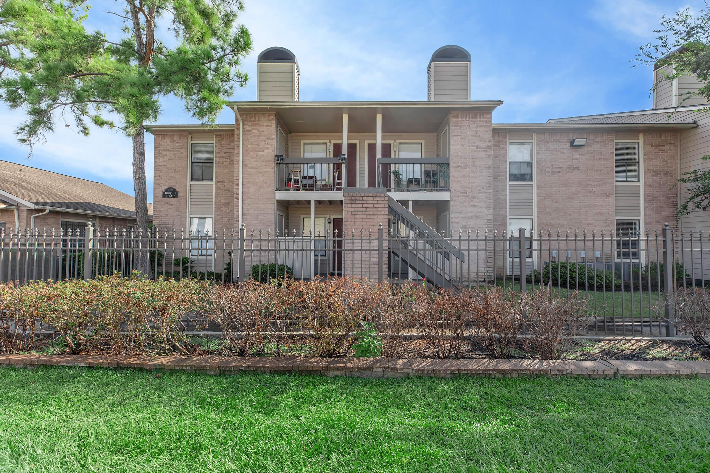 A multi-unit apartment building with a light brick exterior, featuring two upper-level units. There are two ventilation units on top, a staircase leading to the balcony, and a fenced front yard with neatly trimmed shrubs and grass. Trees are visible nearby under a clear blue sky.