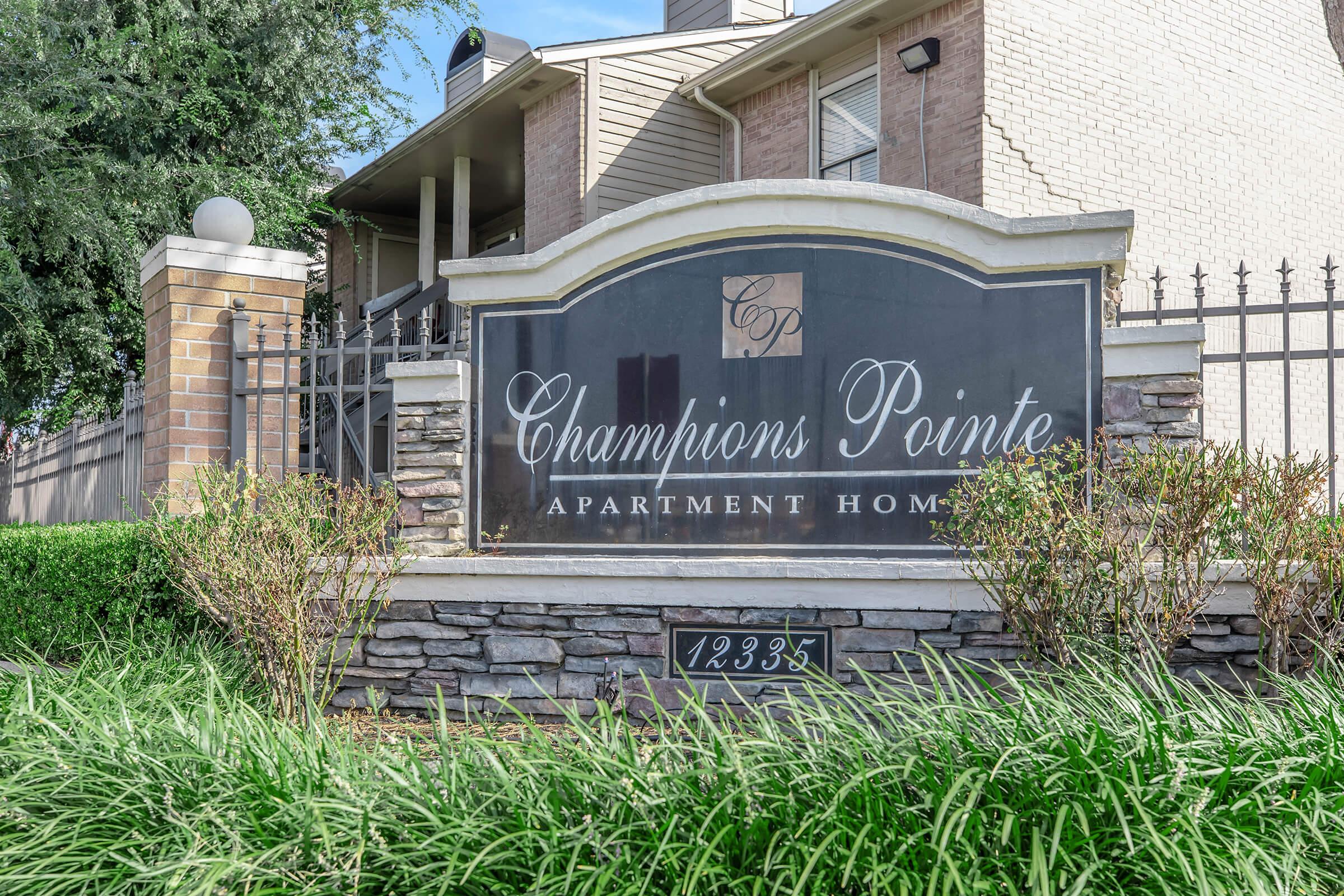 Sign for Champions Pointe Apartment Homes, featuring dark stone and light brick elements, surrounded by greenery. The address displayed is 12335. The entrance is marked with a decorative fence and well-maintained landscaping, emphasizing a welcoming atmosphere for residents.