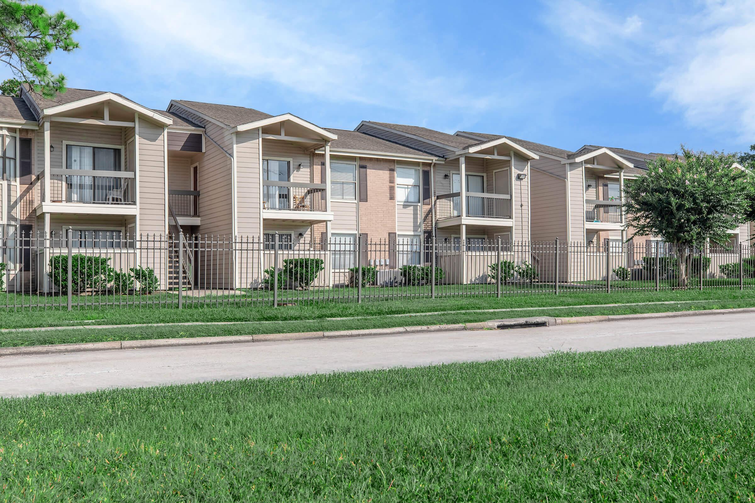 Two-story apartment buildings with balconies, surrounded by green grass and small shrubs. A fence lines the property, and a paved road runs in front of the buildings under a clear blue sky.