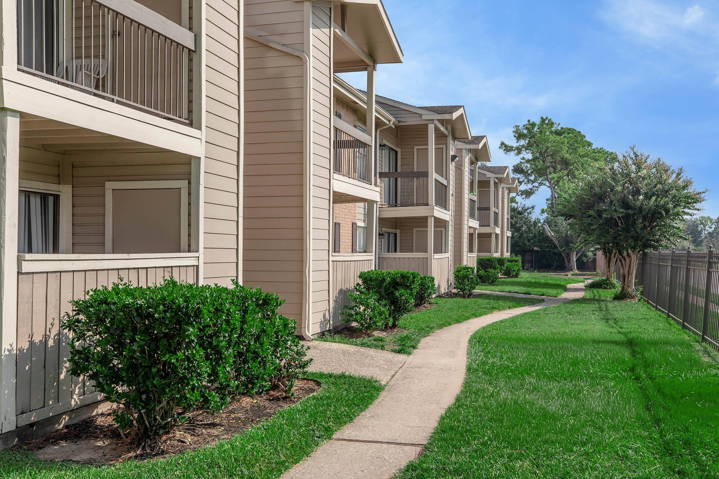A view of a residential apartment complex featuring multiple units with light-colored siding, balconies, and well-maintained landscaping. A paved walkway curves through manicured lawns and shrubbery, leading alongside the buildings. The sky is blue with a few clouds, indicating a pleasant day.