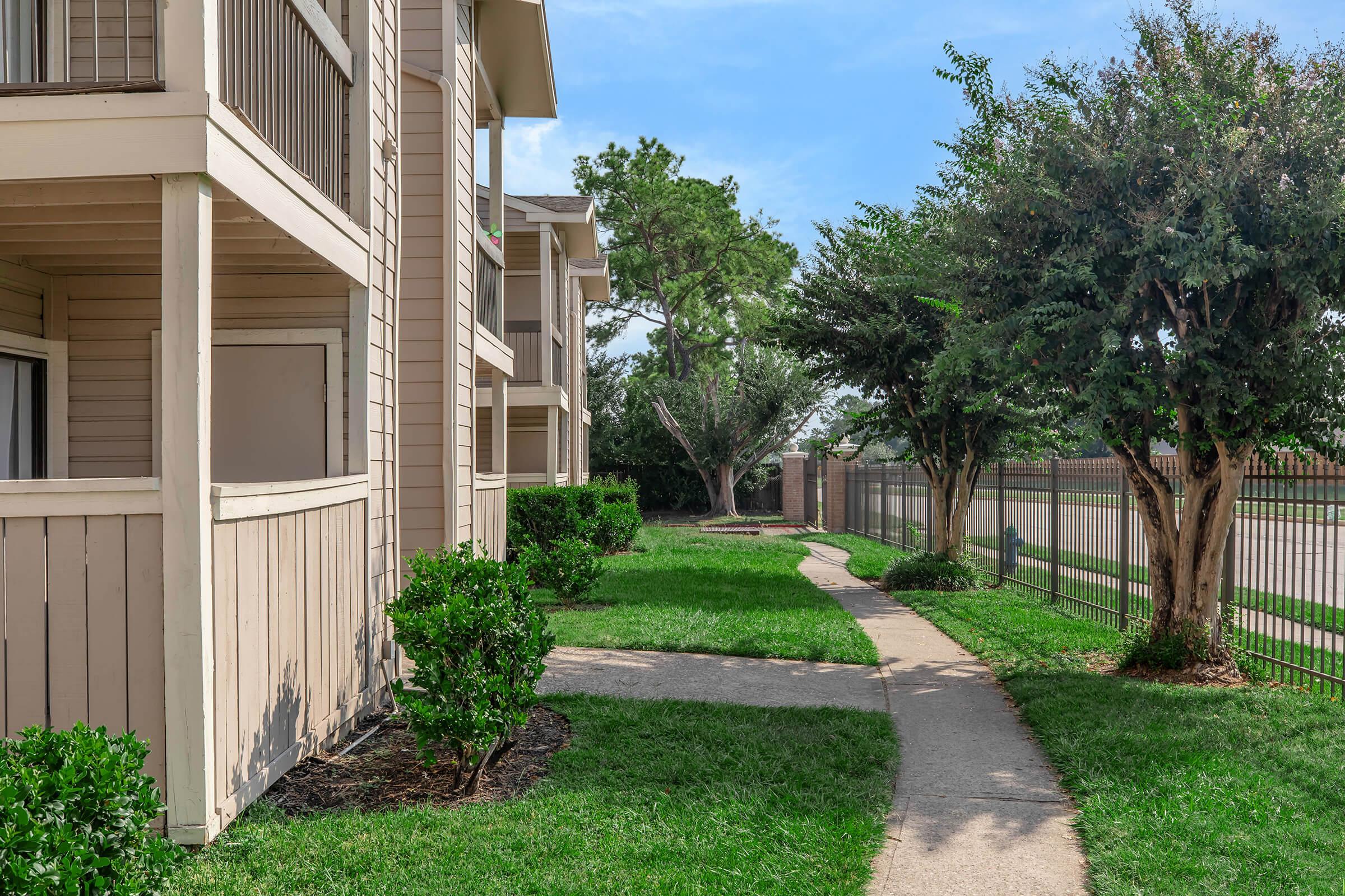 A pathway through well-kept green lawns and plants leads between beige apartment buildings. There are trees and a fence in the background, creating a serene residential scene under a blue sky.