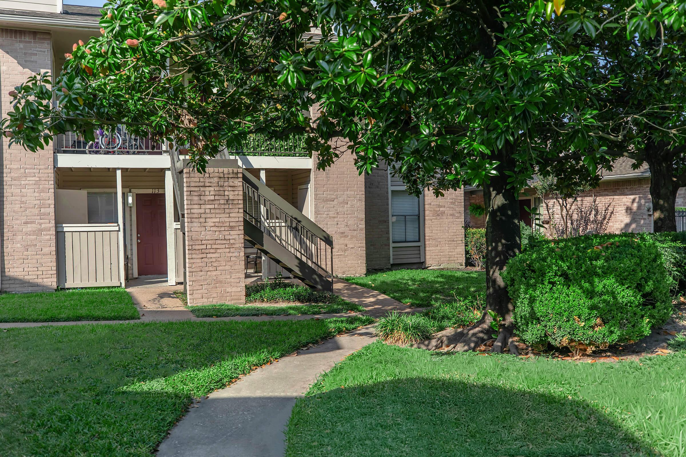 A view of a landscaped apartment complex featuring a brick building with balconies, a set of stairs leading to an upper floor, well-maintained green lawns, and trees. Pathways are visible, and there are hedges and shrubs around the area, contributing to a neatly arranged outdoor environment.