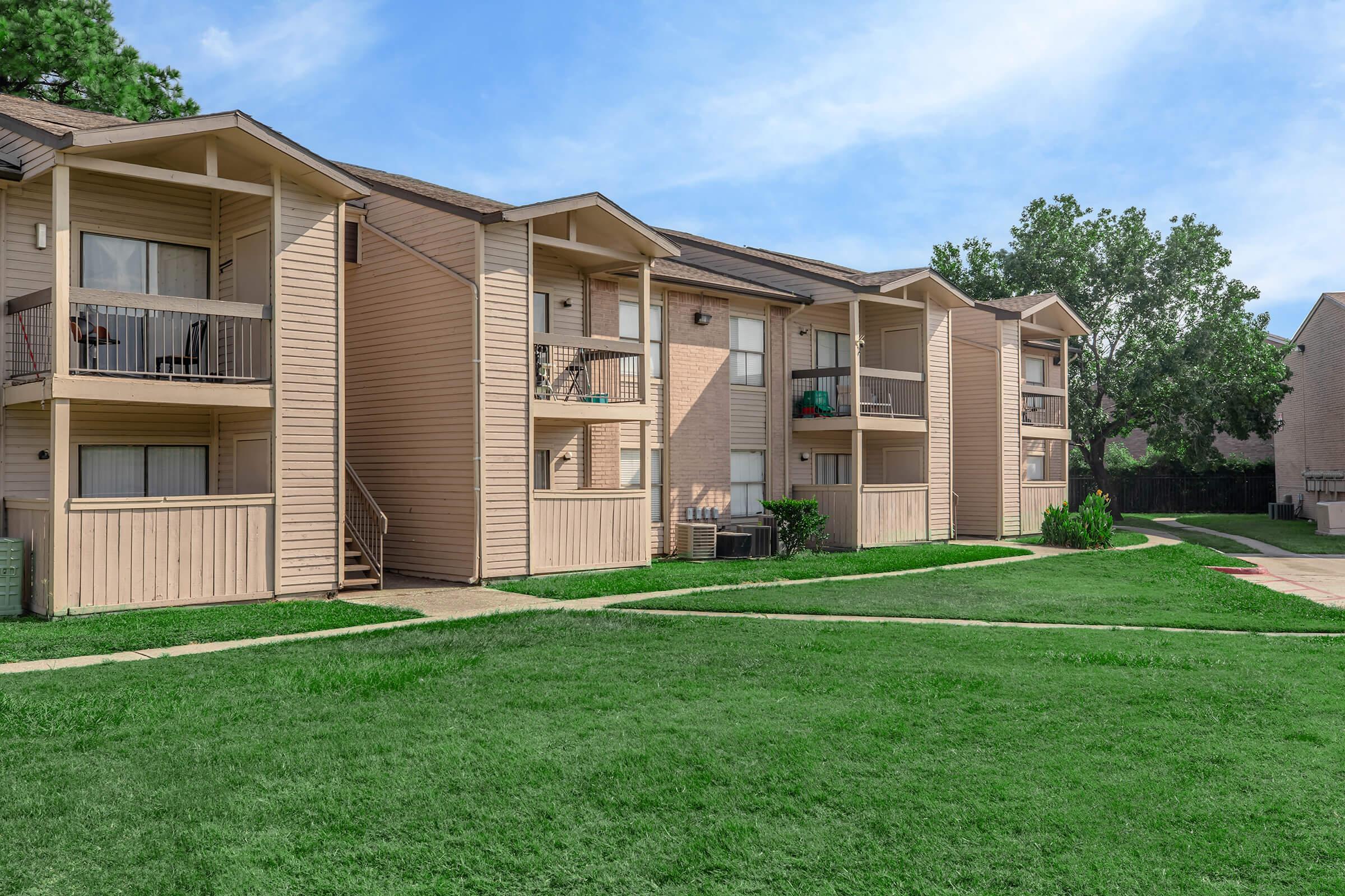 A row of three residential apartment buildings with balconies, surrounded by well-maintained green grass. The buildings are light-colored with wooden siding, and there are pathways leading to each entrance. Trees are visible in the background under a clear blue sky.