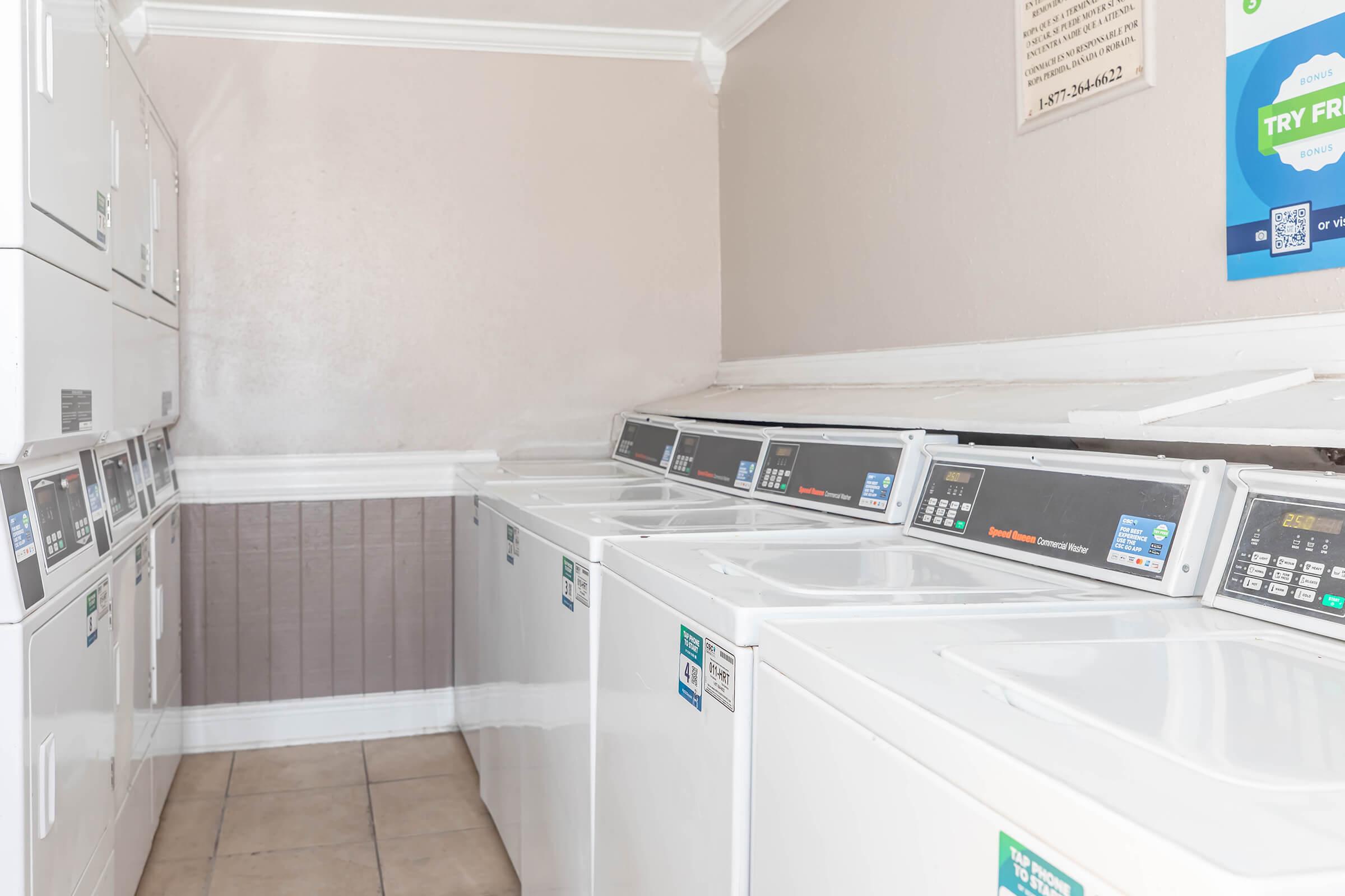 A clean laundry room featuring several stacked washing machines and dryers lined against a light-colored wall. The machines have digital display panels and are in good condition, with a tiled floor. There is a sign on the wall, and the overall atmosphere is bright and tidy.