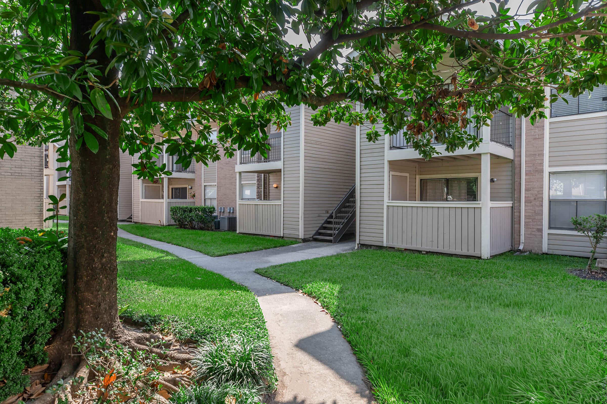 A well-maintained residential area featuring a grassy pathway, surrounded by manicured shrubs and trees. The scene includes two-story apartment buildings with balconies, framed by green foliage, creating a pleasant outdoor living environment. Sunlight filters through the leaves, enhancing the inviting atmosphere.