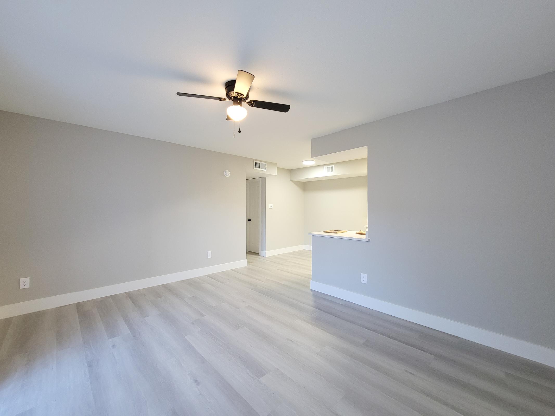 A spacious living area with light gray walls, modern laminate flooring, and a ceiling fan. In the background, there is a doorway leading to another room and a hint of a kitchen area with an unobtrusive bar counter. Natural light fills the space, enhancing its fresh and bright atmosphere.