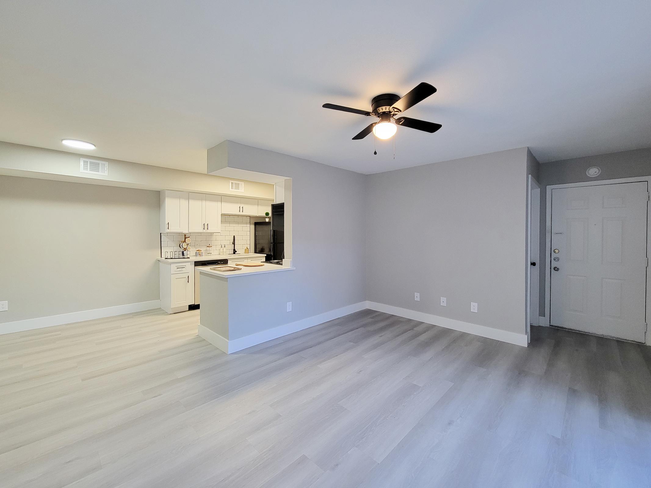 A spacious living area with light-colored walls and grey laminate flooring. A ceiling fan is installed, and a small kitchen area is visible through an opening, featuring white cabinets and a countertop. The entrance door is shown on the right side of the image.