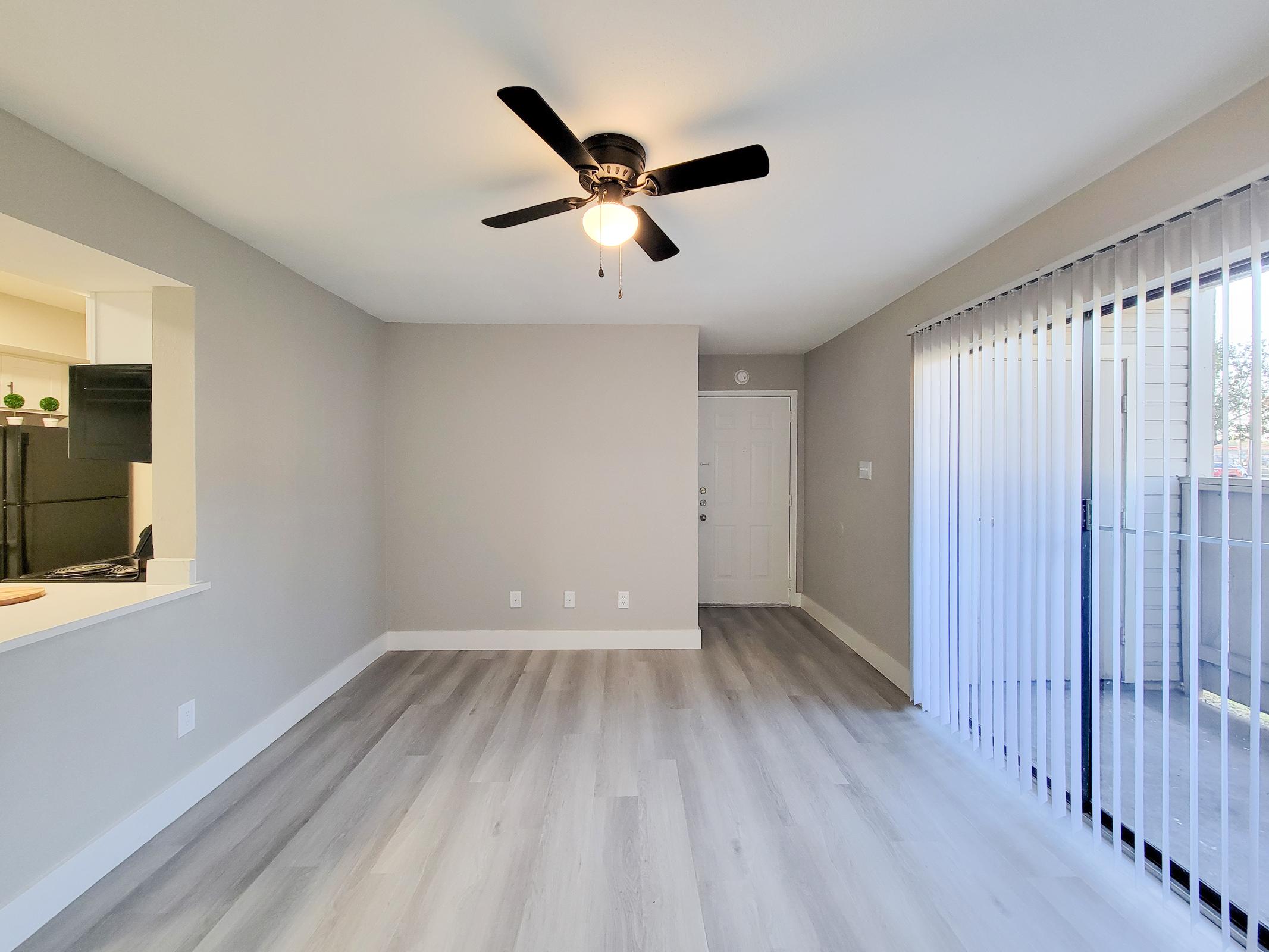 A bright, empty living room featuring light gray walls and sleek vinyl flooring. A ceiling fan is mounted above, and large sliding glass doors offer access to an outdoor space. On the left, there is an open kitchen area with minimal decor, creating a spacious and airy atmosphere.