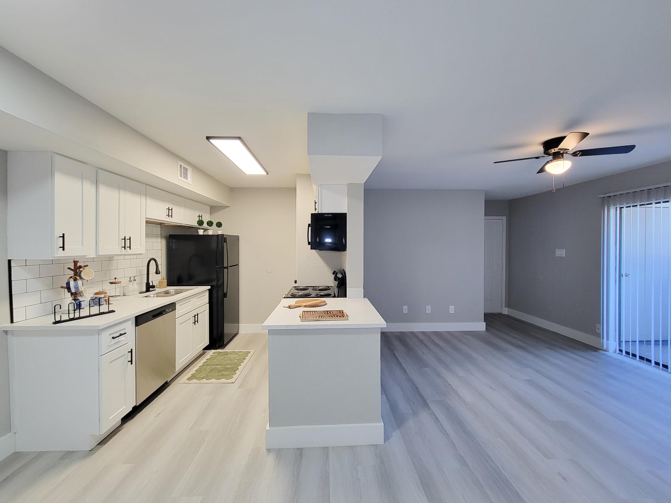 A modern kitchen featuring white cabinets, a stainless steel sink, and a black refrigerator. The kitchen island has a light countertop and is open to a spacious living area with a ceiling fan and large windows. The flooring is light-colored and the walls are painted in a neutral tone.