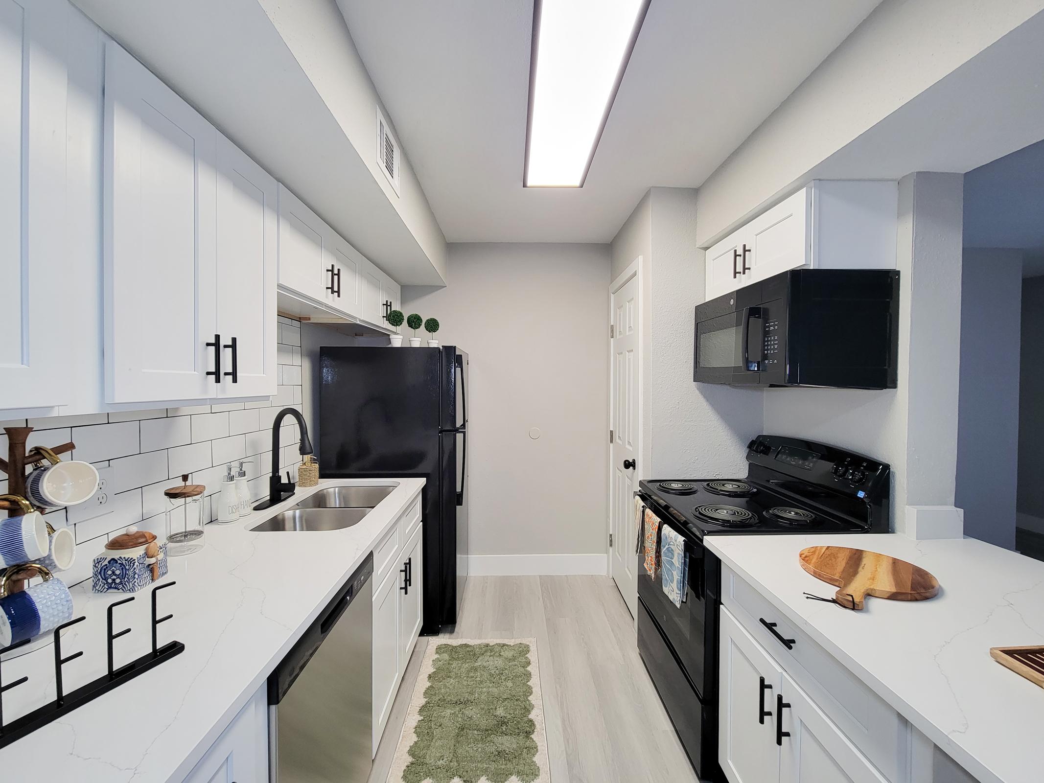 Modern kitchen scene featuring white cabinetry, a dark refrigerator, and a black stove. The countertops are sleek and light-colored, with a decorative cutting board and kitchenware displayed. Natural light from the overhead fixture brightens the space, which includes a green rug on the floor, creating a fresh and inviting atmosphere.