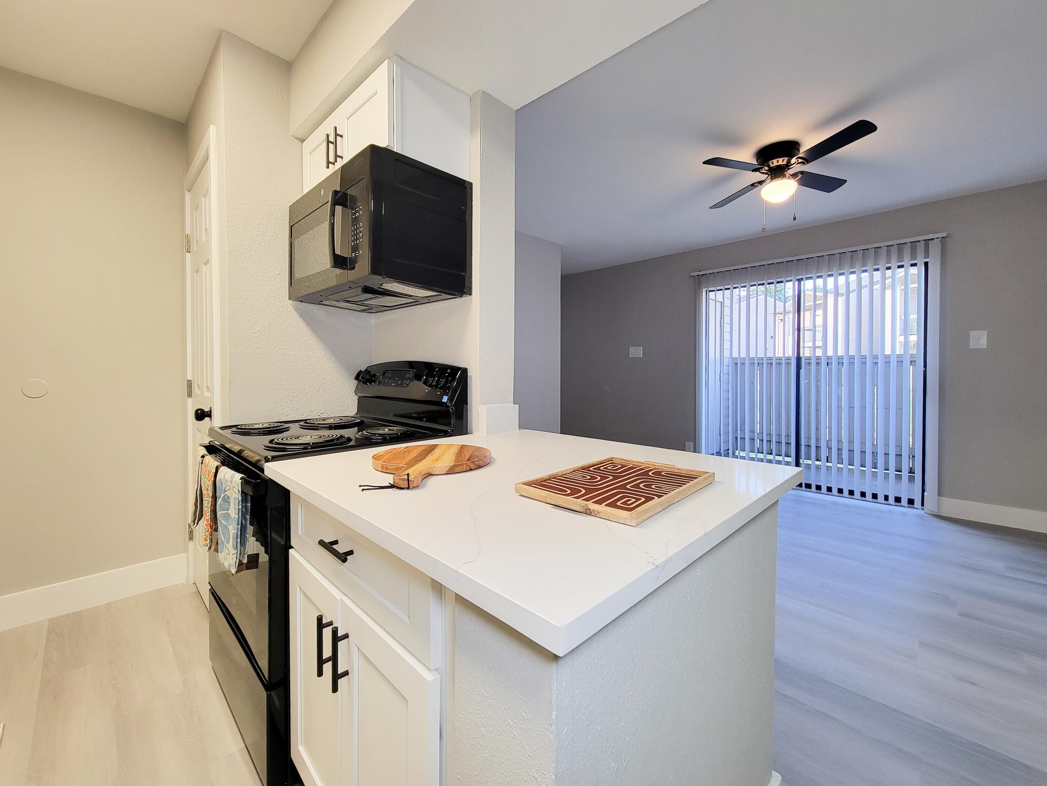 A modern kitchen with a black stove, microwave, and white cabinetry. A wooden cutting board sits on the countertop, and the open living area features a ceiling fan and sliding glass doors leading to an outdoor space. Light-colored flooring enhances the bright, airy atmosphere.