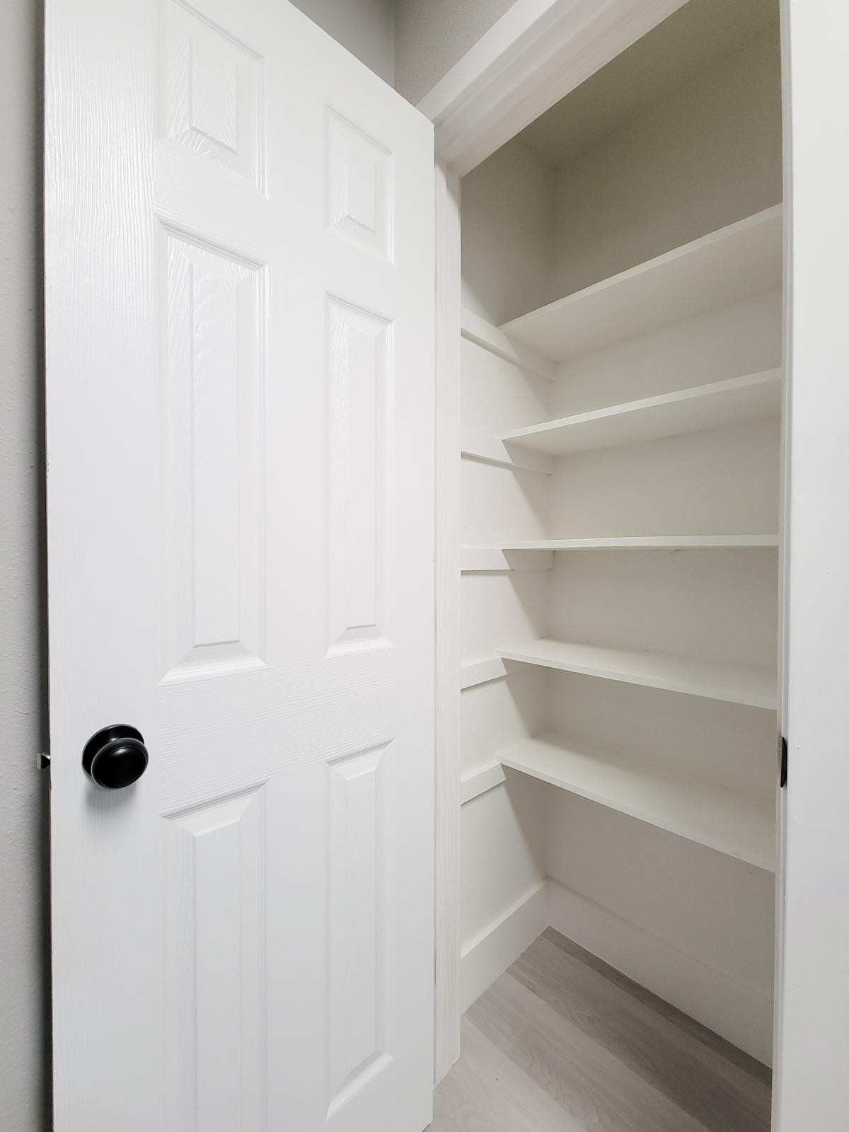 A view of an open white door leading into a clean, empty pantry with three shelves against a light-colored wall. The floor has a light finish, and the overall space is well-lit and minimalistic.