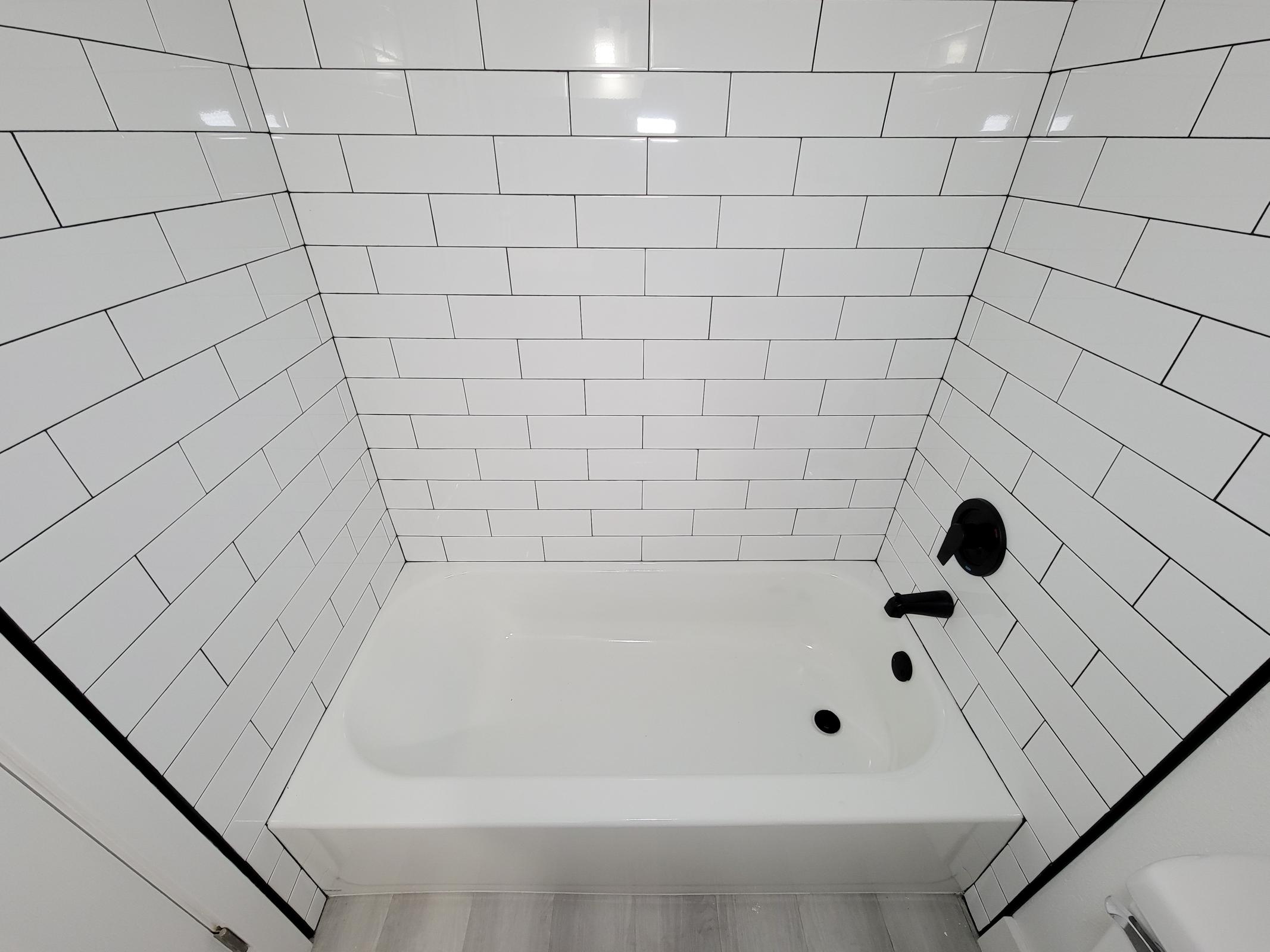 A clean, modern bathroom with a white tiled wall featuring black grout. The bathtub is white and positioned against the tiled wall, with a black faucet and shower fixture. The floor is light-colored, complementing the overall design of the space.
