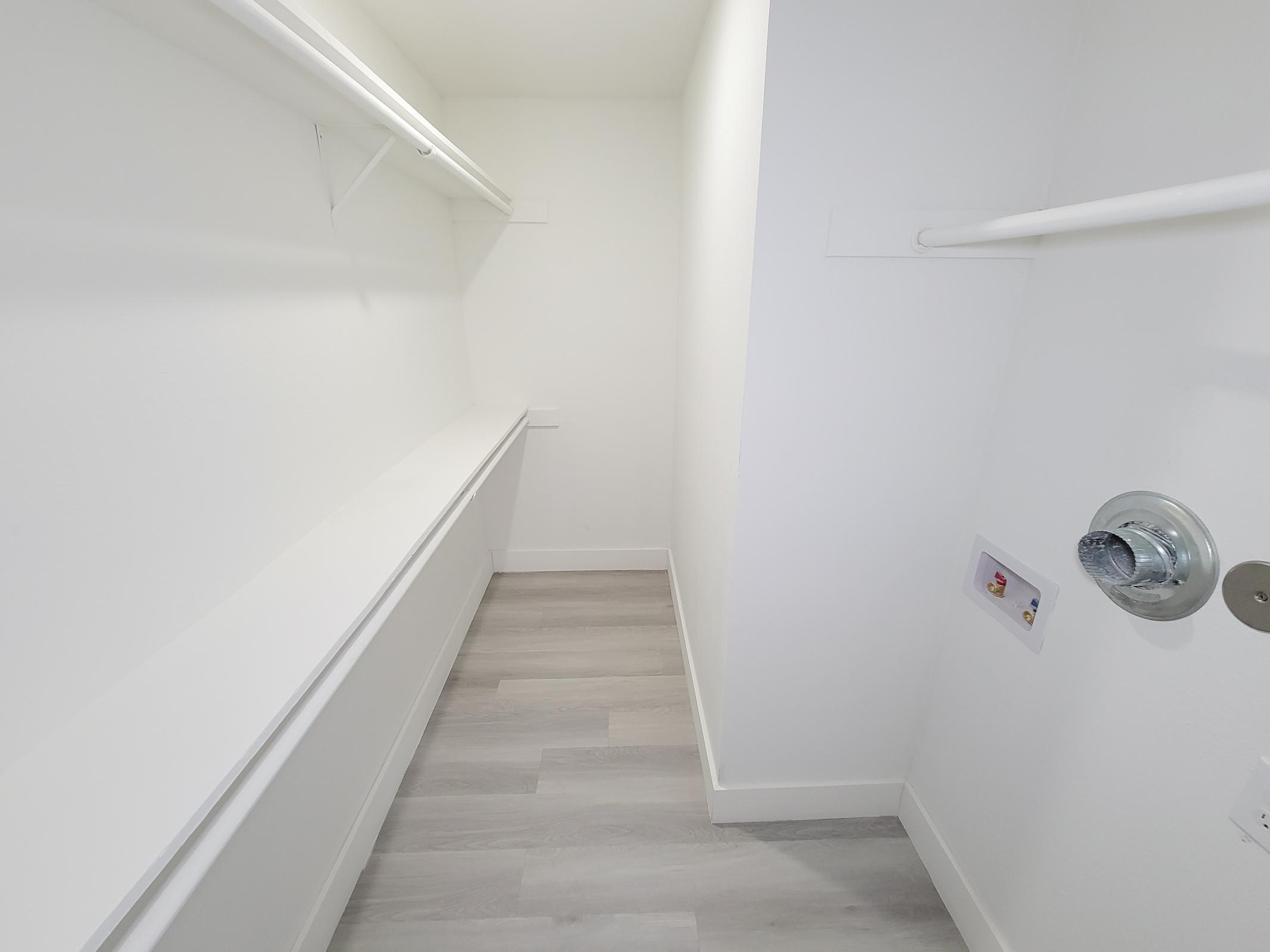 A spacious, empty closet featuring light gray walls and a light gray floor. It has two white shelves along the sides and a wall-mounted electrical outlet. The closet is well-lit, with a clean and minimalistic design.