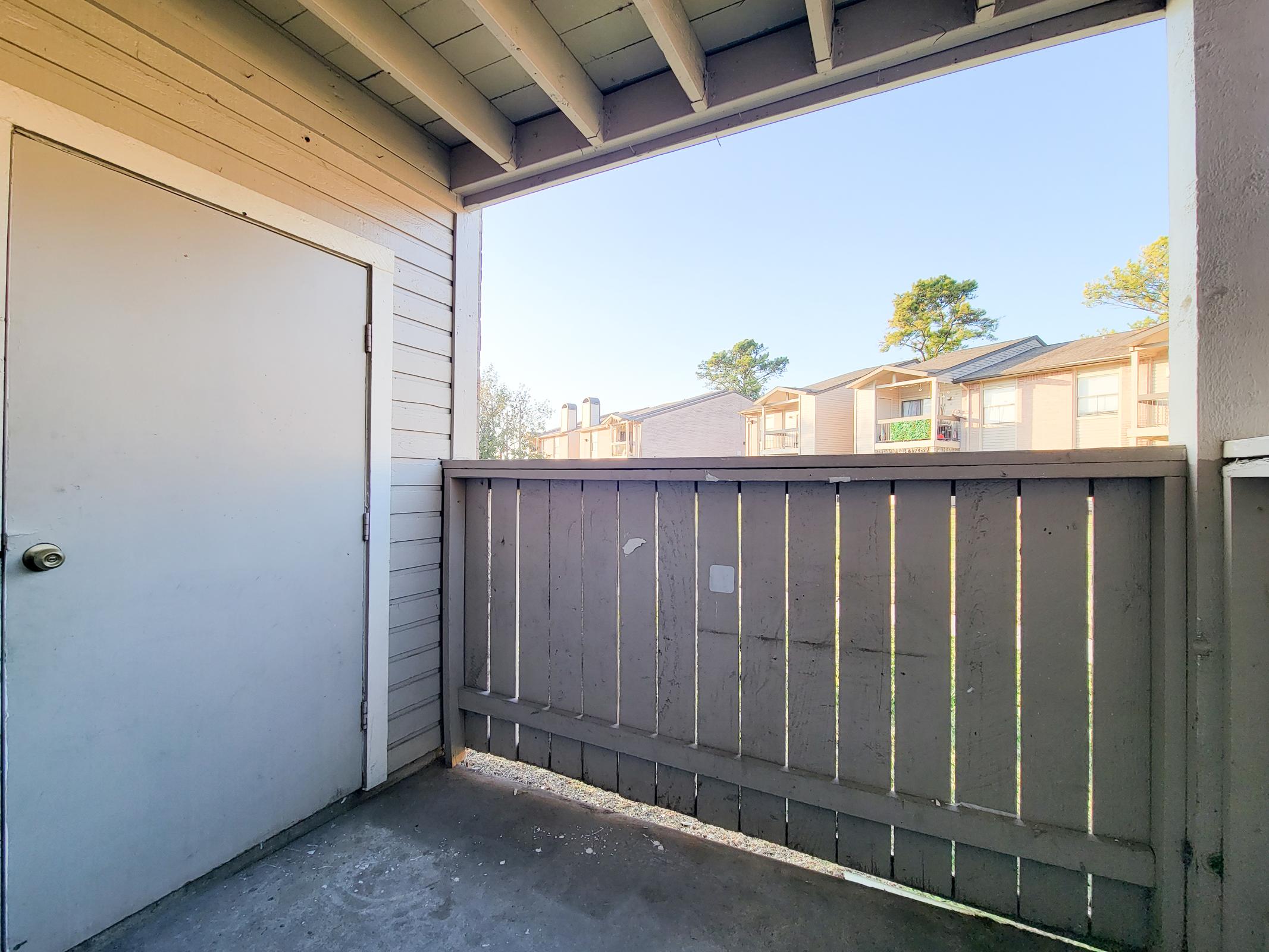 View from a balcony showing a closed door and a wooden railing. The scene features a clear blue sky and buildings in the distance, hinting at an outdoor space in an apartment complex.