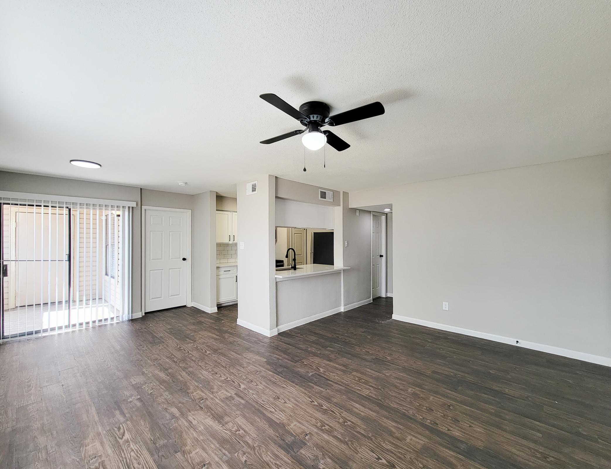 Spacious interior of an apartment featuring a living area with wooden flooring, a ceiling fan, and a kitchen area in the background. Natural light enters through sliding glass doors. Freshly painted walls give a modern and clean look.