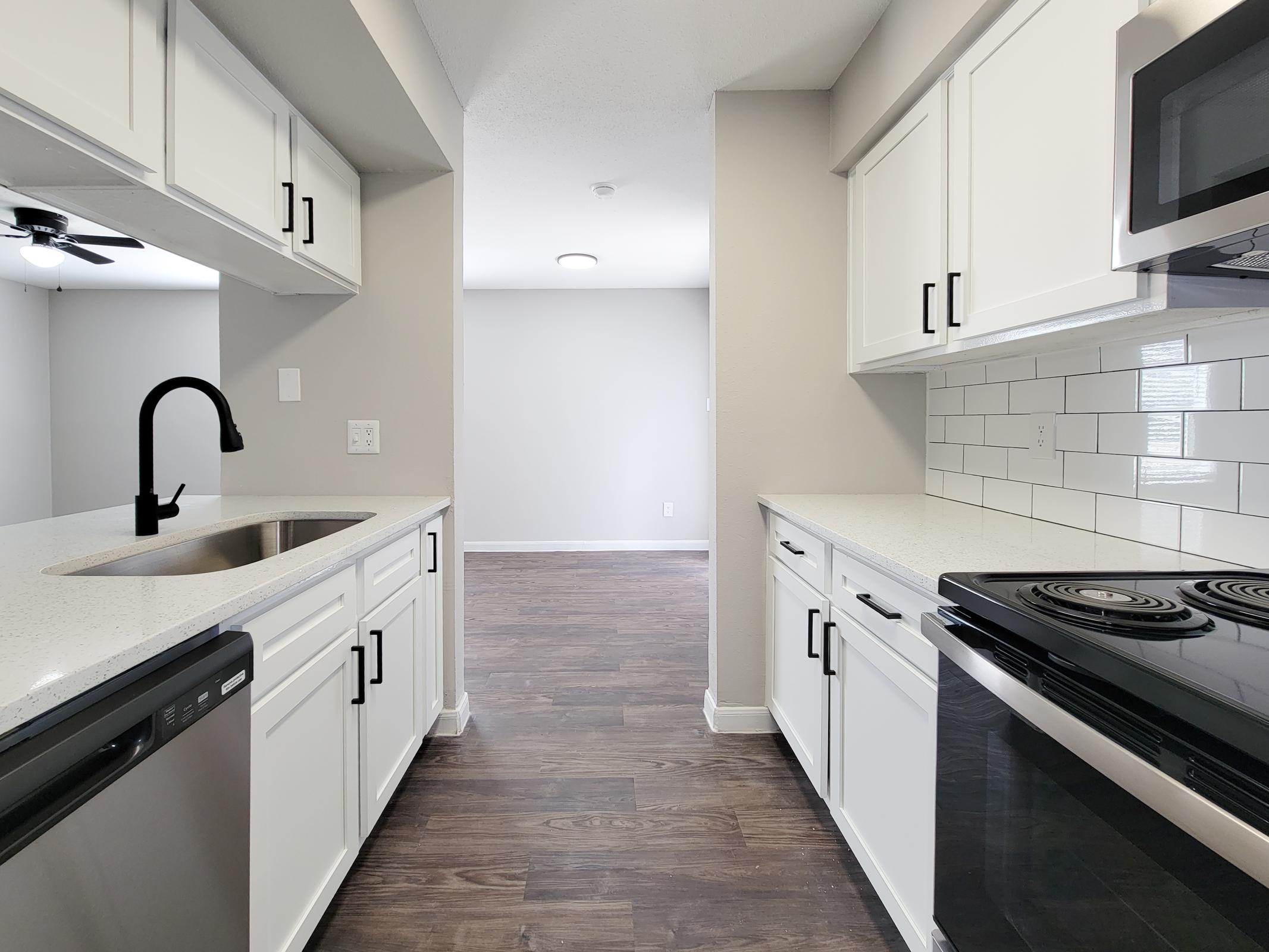 Modern kitchen with white cabinetry, black faucet, and stainless steel appliances. Features a spacious countertop and tiled backsplash. Light streaming in from a nearby window creates a bright and inviting atmosphere. In the background, an open living area with light gray walls is visible.