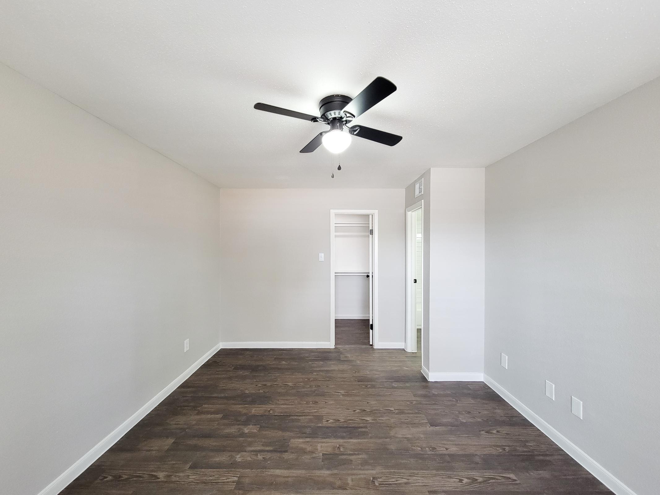 Bright, spacious room with light gray walls and dark wood flooring. A ceiling fan with black blades is installed in the center. A closet with white shelving is visible in the background, along with a door leading to another space. The overall atmosphere is modern and inviting.