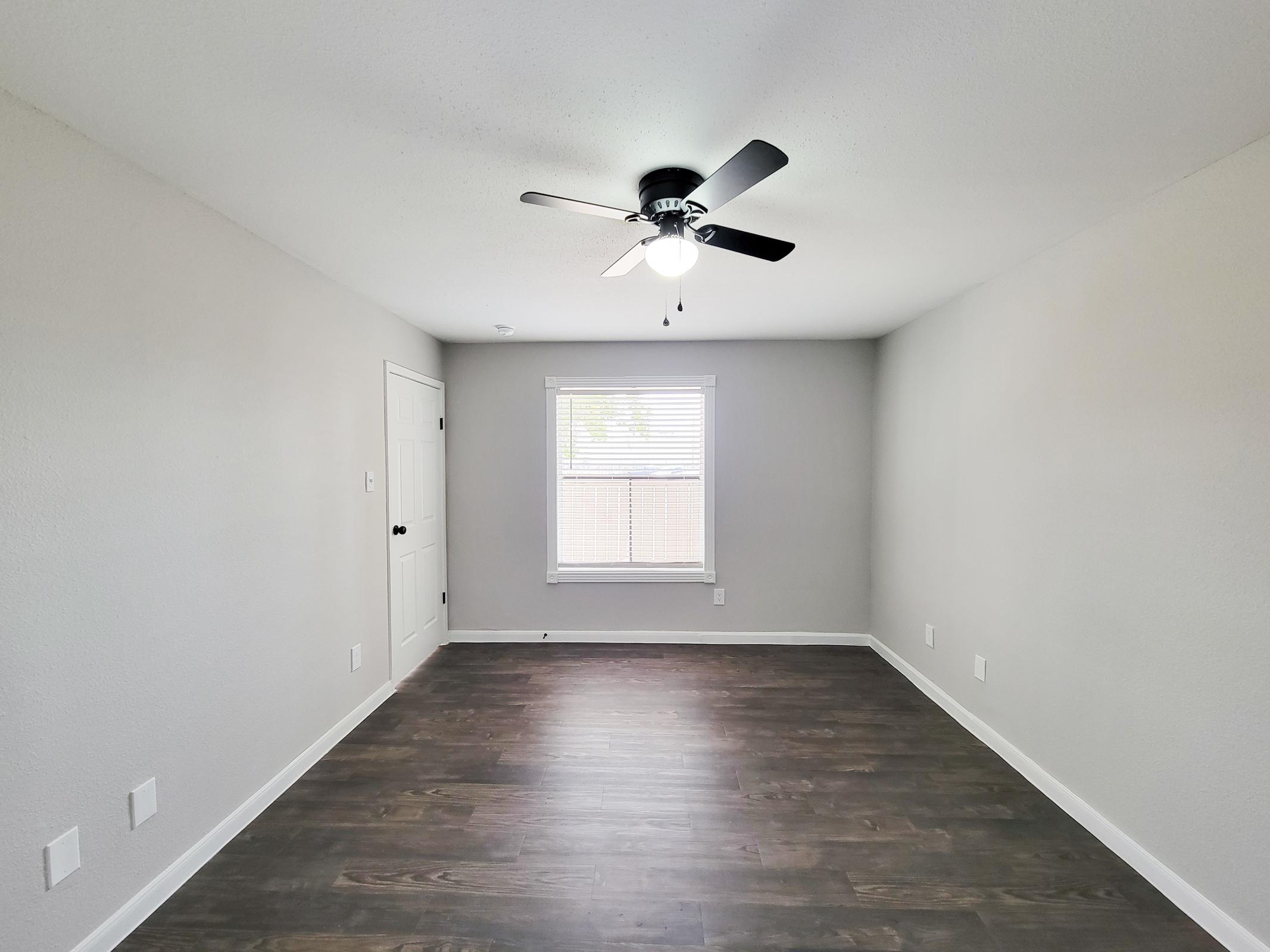 Empty room with light gray walls and a dark wood floor. A ceiling fan with a light fixture hangs in the center. There is a small window with blinds allowing natural light, and a single door on one wall. The overall space appears clean and modern, suitable for various uses.