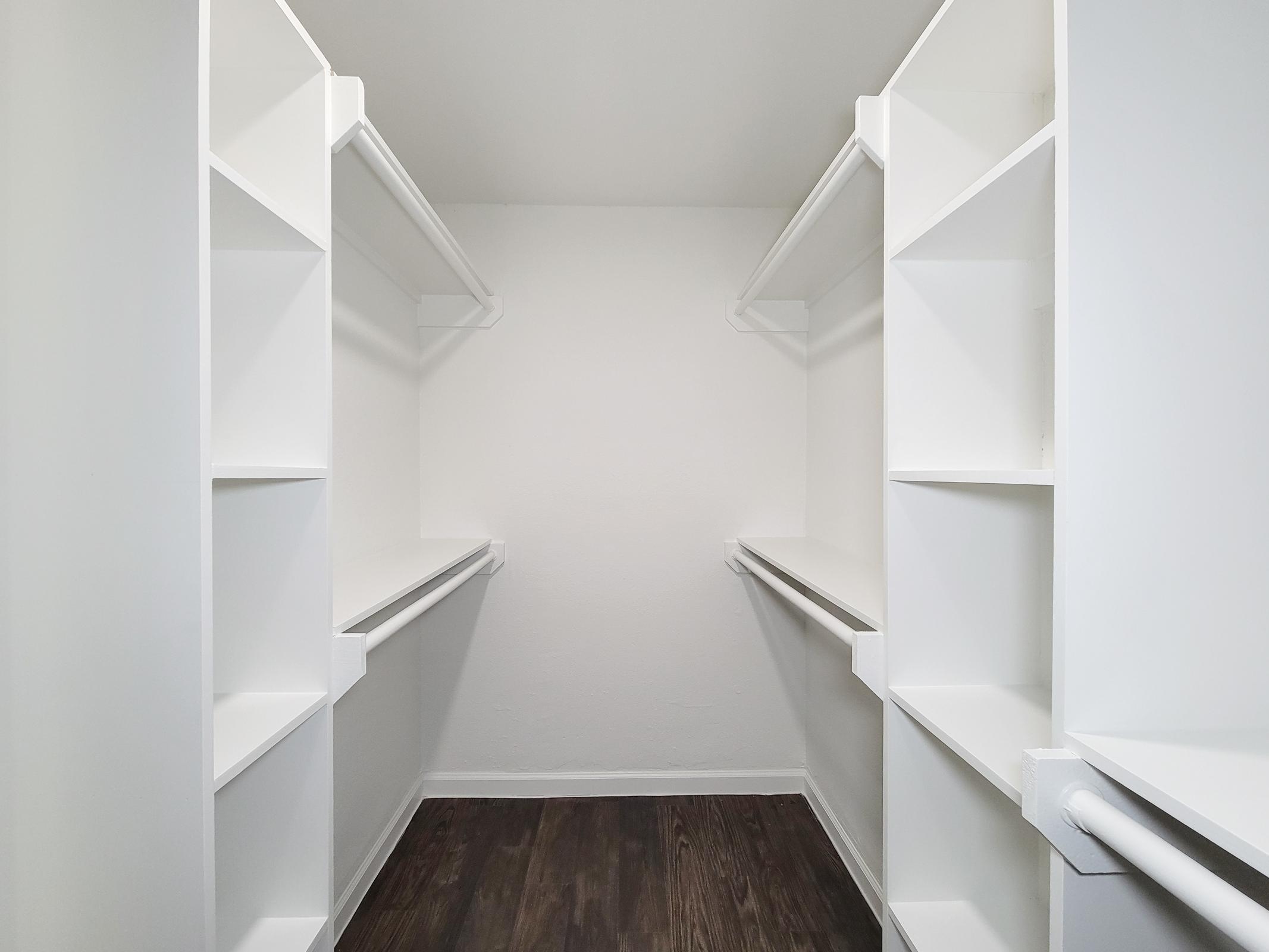 A spacious walk-in closet featuring white shelving units and hanging rods on either side, with a neutral wall color and dark wooden flooring, creating a clean and organized storage space.
