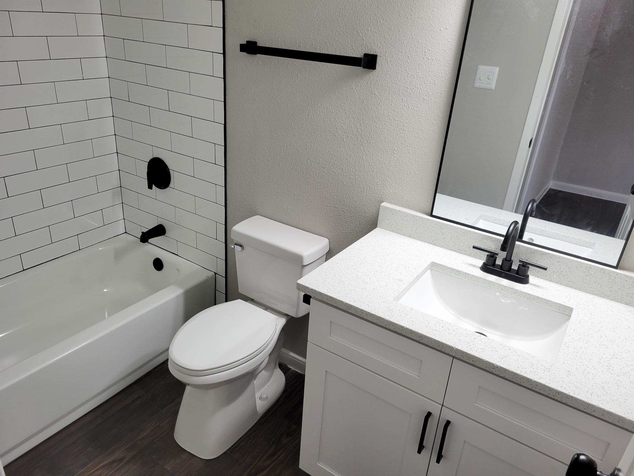 A modern bathroom featuring a bathtub with white tiled walls, a black faucet, a white toilet, and a white countertop with a sink. The room has a neutral color scheme and contemporary fixtures, including a black towel bar and a large mirror above the sink.