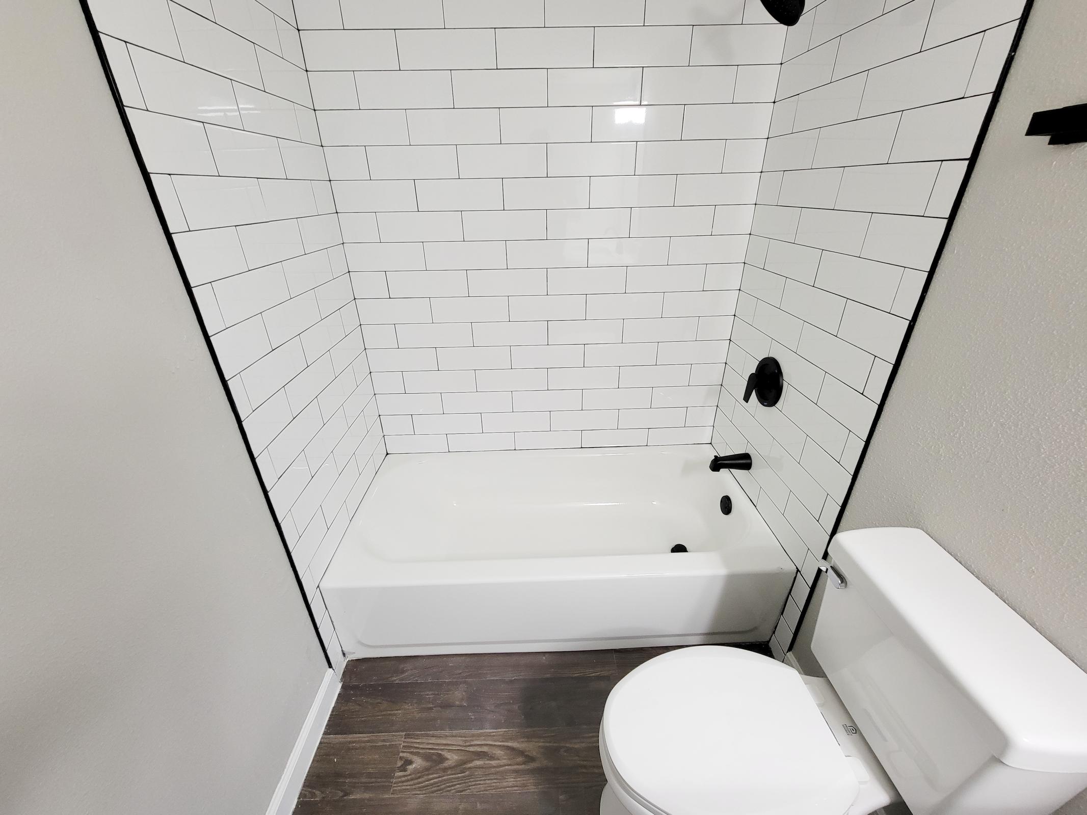 A clean bathroom featuring a white tiled shower with a bathtub, black fixtures, and a white toilet. The floor is a wood-like laminate, creating a modern and minimalist aesthetic.