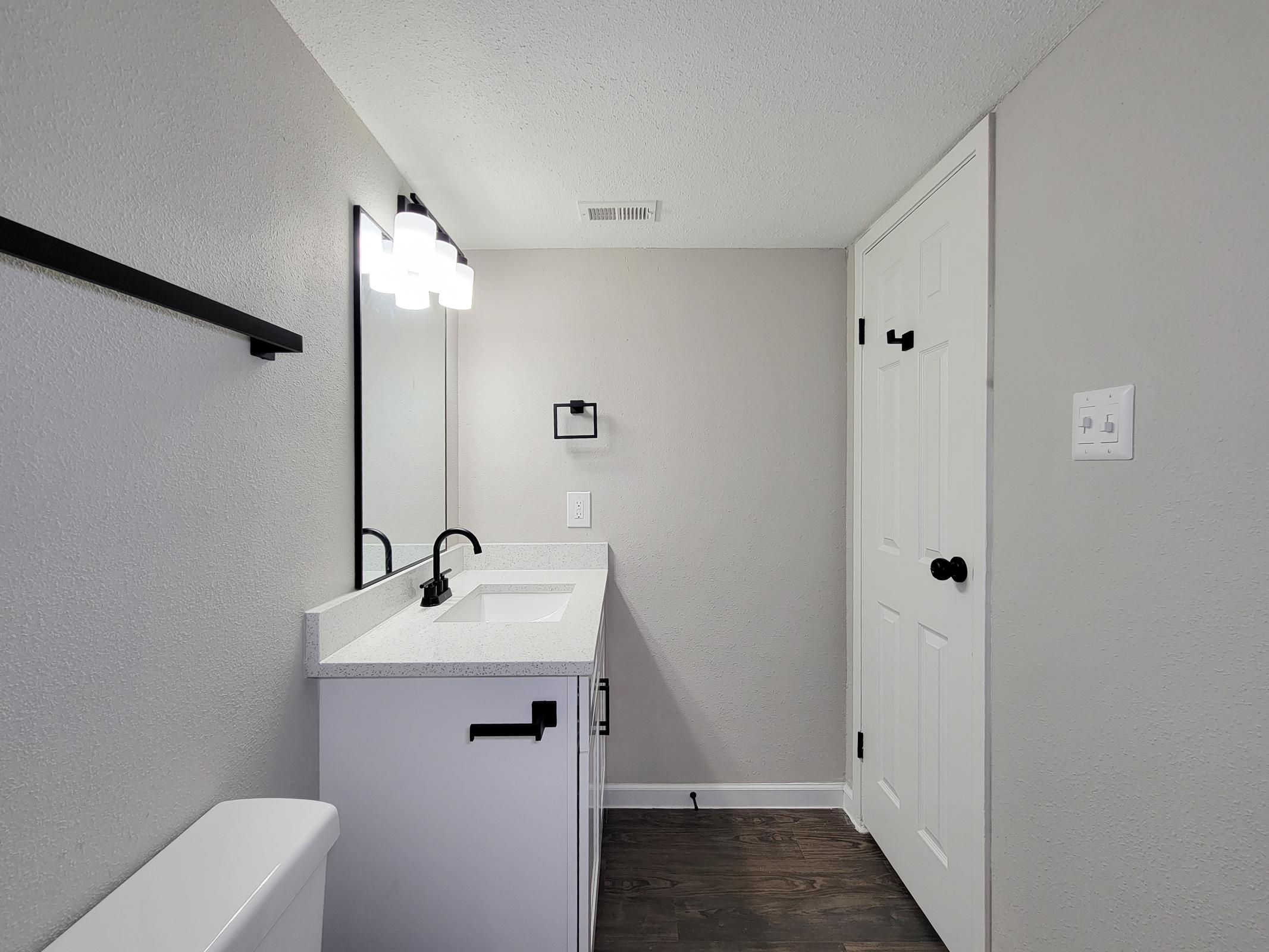 A modern bathroom interior featuring a white sink with a countertop, a wall-mounted mirror, and stylish lighting fixtures. The walls are light gray, and there’s a black towel rack. A white toilet is visible, along with a closed door and a light switch. The flooring is dark wood, creating a contrast with the lighter elements.