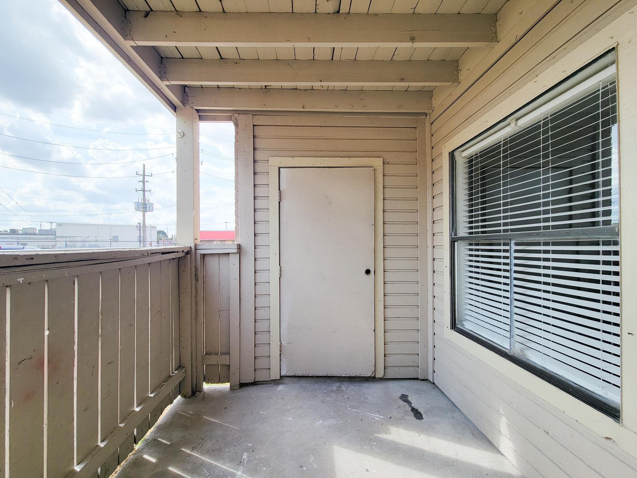 A spacious balcony with a wooden railing, featuring a plain white door on one side and a large window with blinds on the other. The floor is bare and concrete, and there are shadows from the railings. The sky is visible in the background, indicating a bright day.
