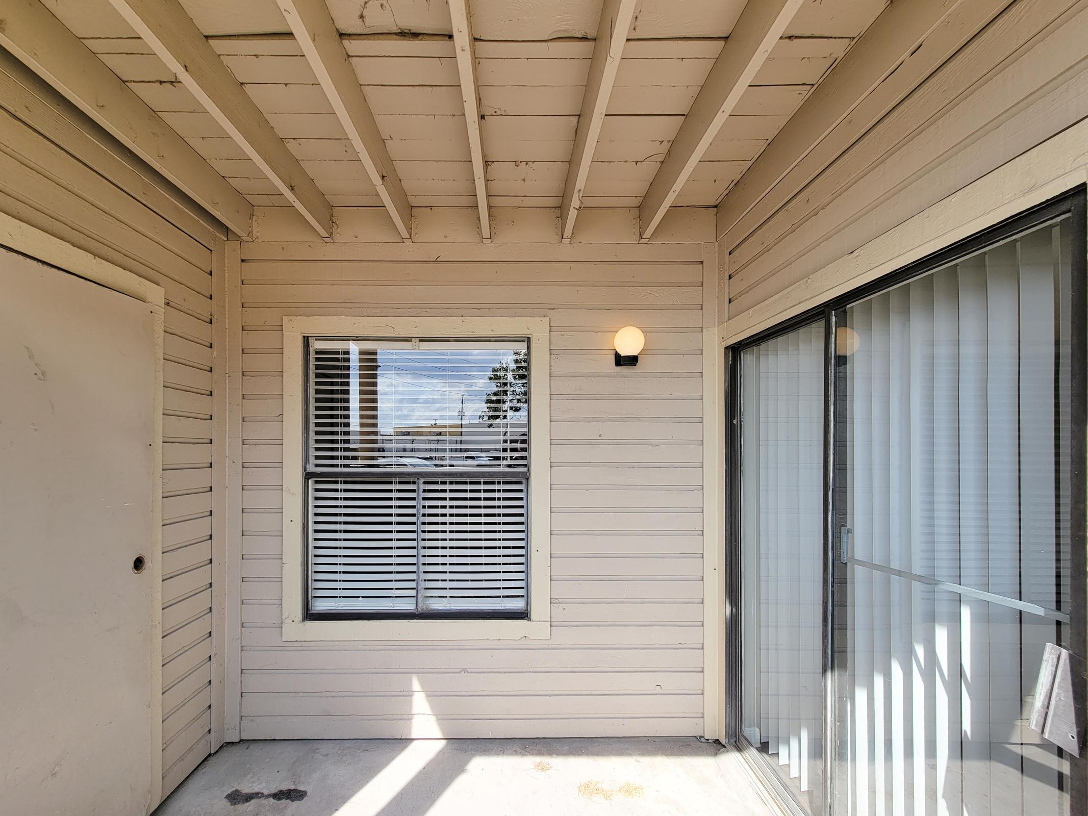 A view of a covered porch area featuring a wooden ceiling, a light fixture on the wall, a large window with white blinds, and a sliding glass door. The space is bright and minimally furnished, with beige wooden siding. Sunlight streams in, creating a warm ambiance.