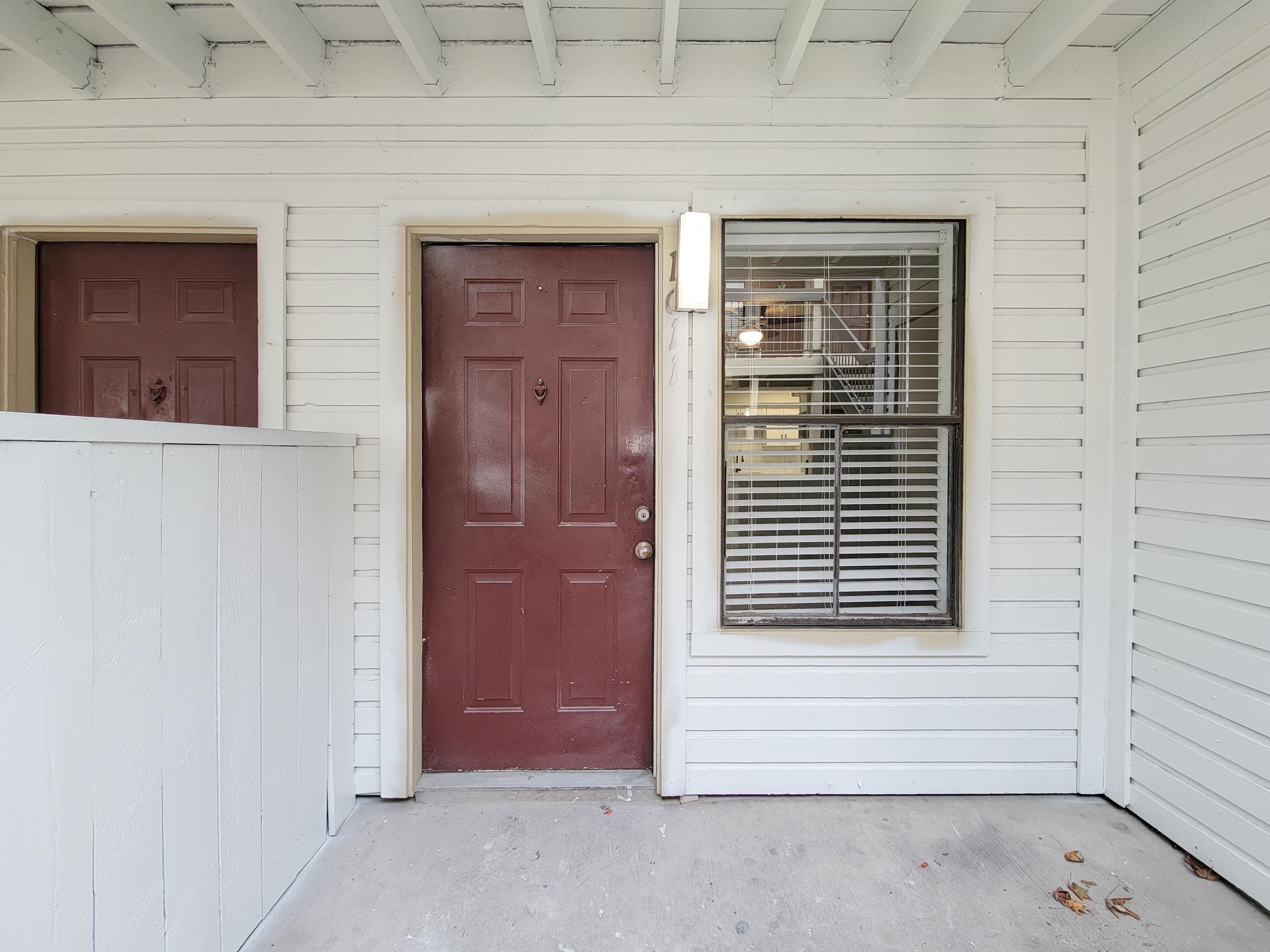 A view of an exterior entrance to an apartment, featuring a maroon front door with a small brass knocker. To the left, there is a white wooden barrier, and to the right, a window with blinds partially drawn. The area is painted white, with visible beams on the ceiling.
