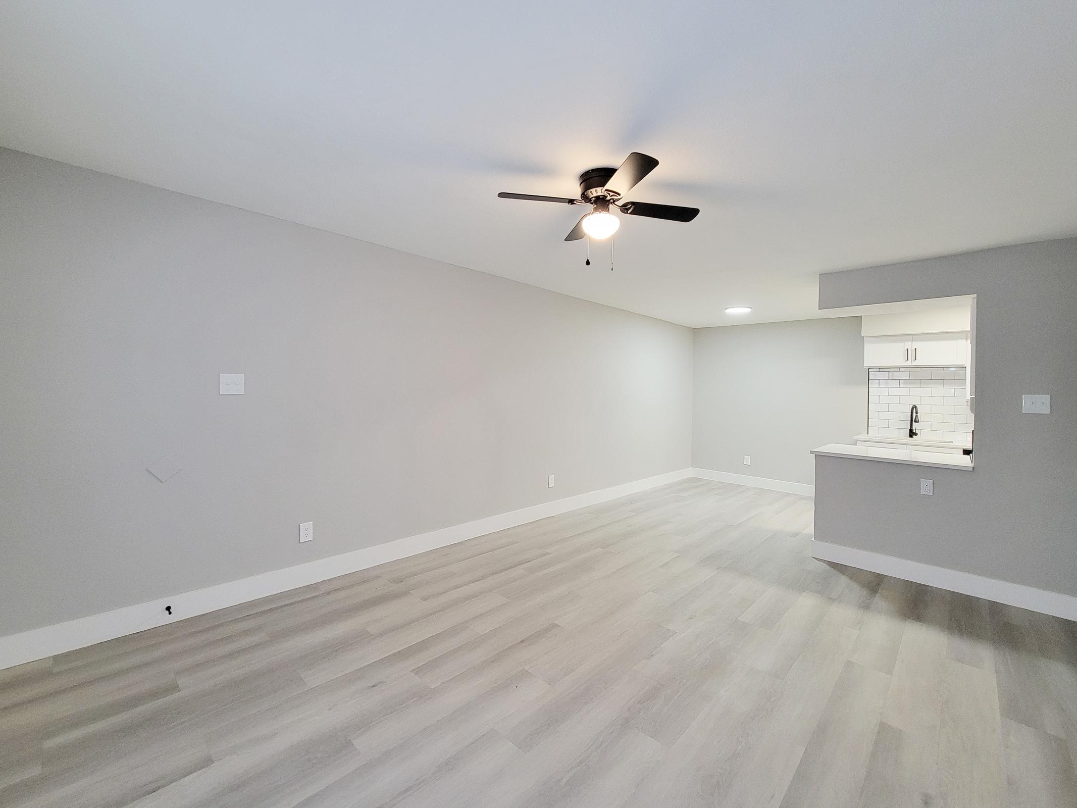 A spacious, modern living room featuring light gray walls and hardwood-style flooring. The room is bright and airy, with a ceiling fan and a pass-through to a kitchen area partially visible in the background. The overall design is contemporary and minimalist.