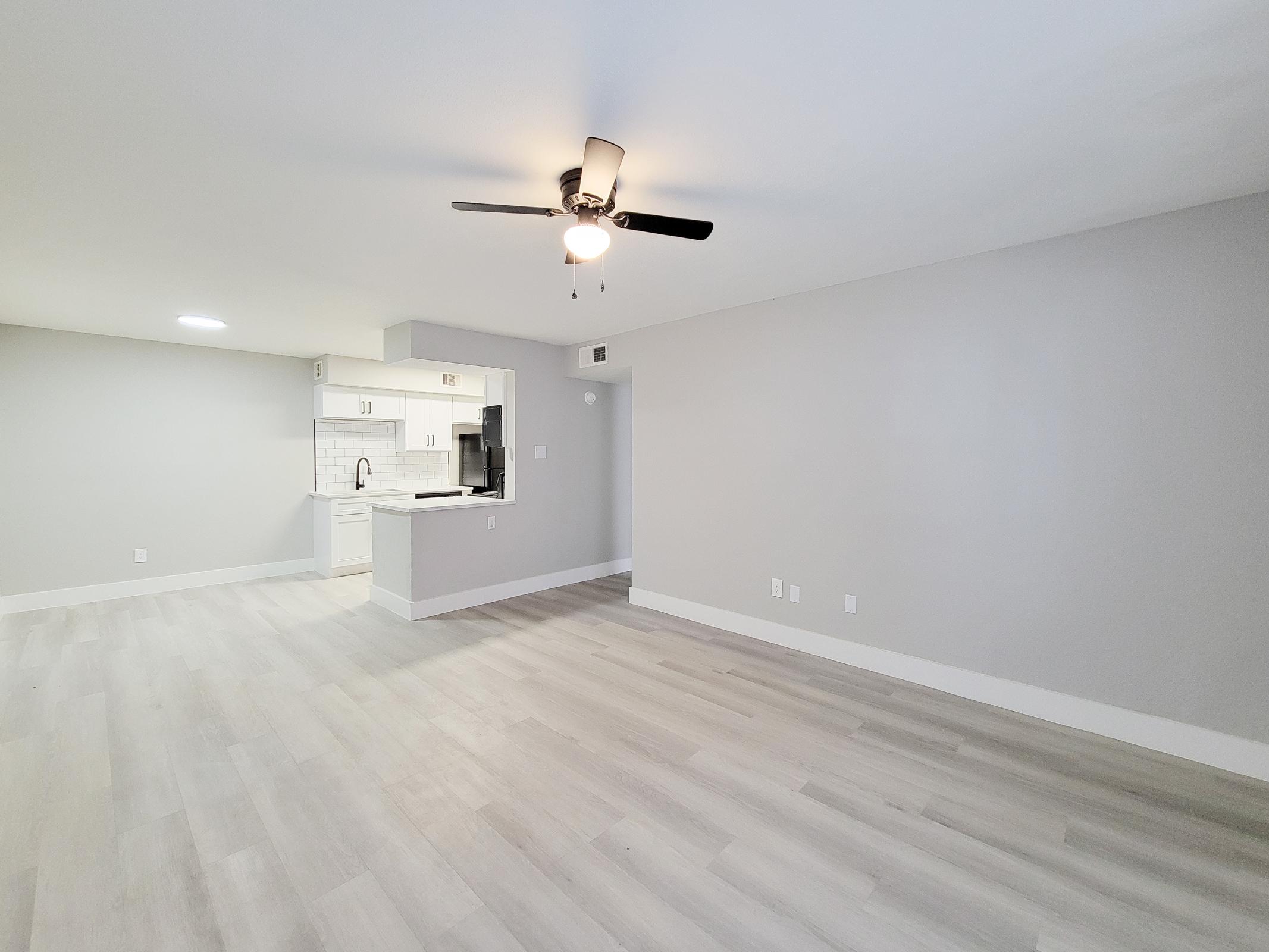 A bright and spacious living area featuring light-colored walls and flooring. A ceiling fan is visible, and a modern kitchen with white cabinetry and countertops is in the background. The layout is open, creating a welcoming atmosphere.