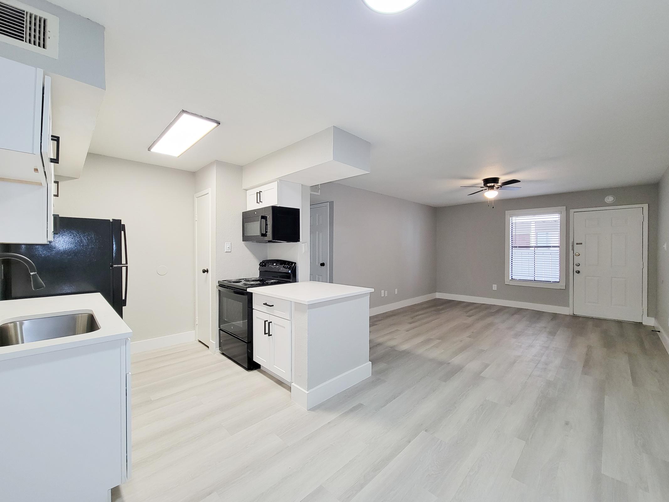 A bright and modern kitchen and living area featuring white cabinetry, a black refrigerator, and stove. The open layout includes a small island with a sink, light-colored flooring, and ample natural light from a window. A ceiling fan is visible in the living area, which also has a front door.